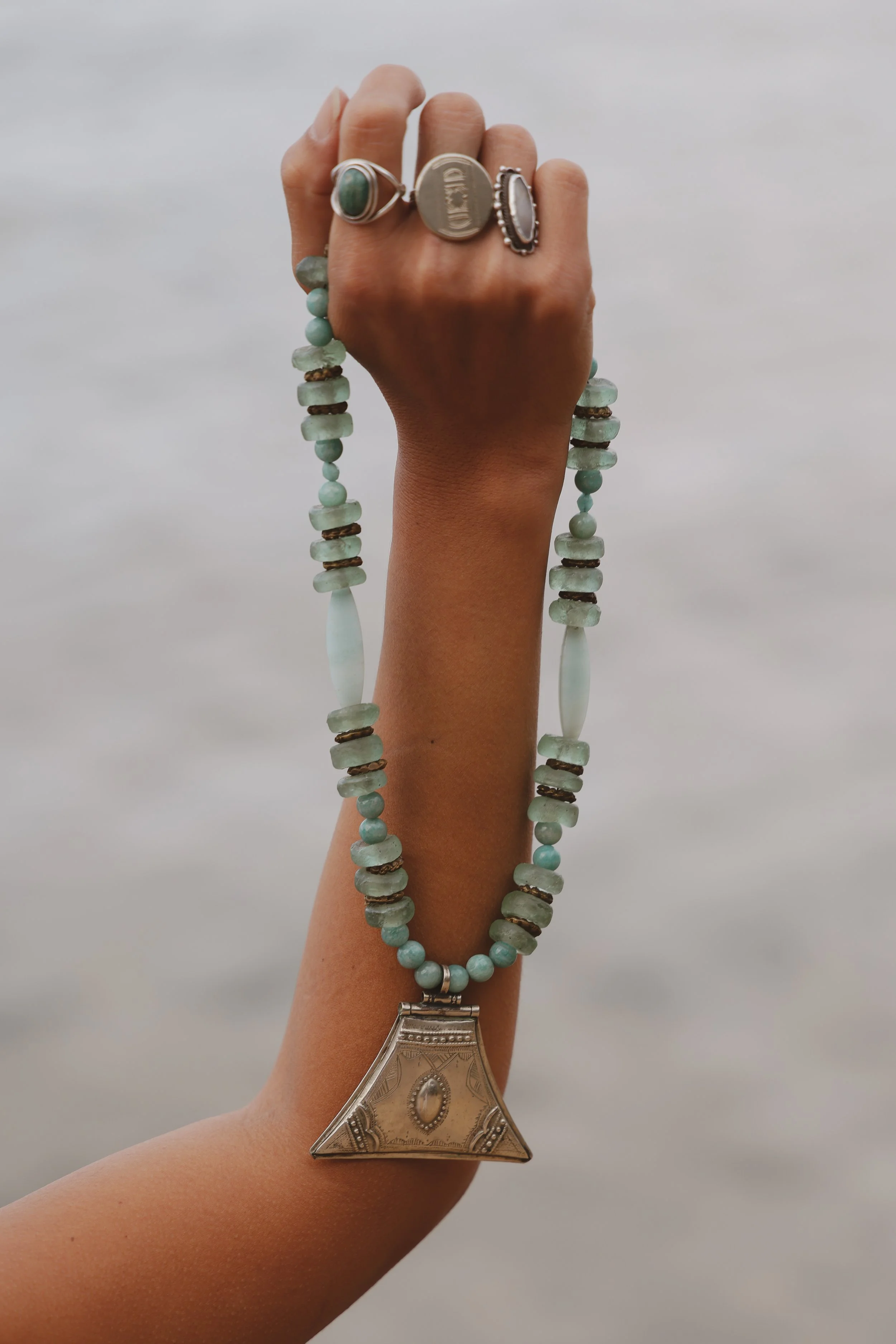 A person's hand holding a beaded necklace with a large silver pendant against a cloudy sky background.