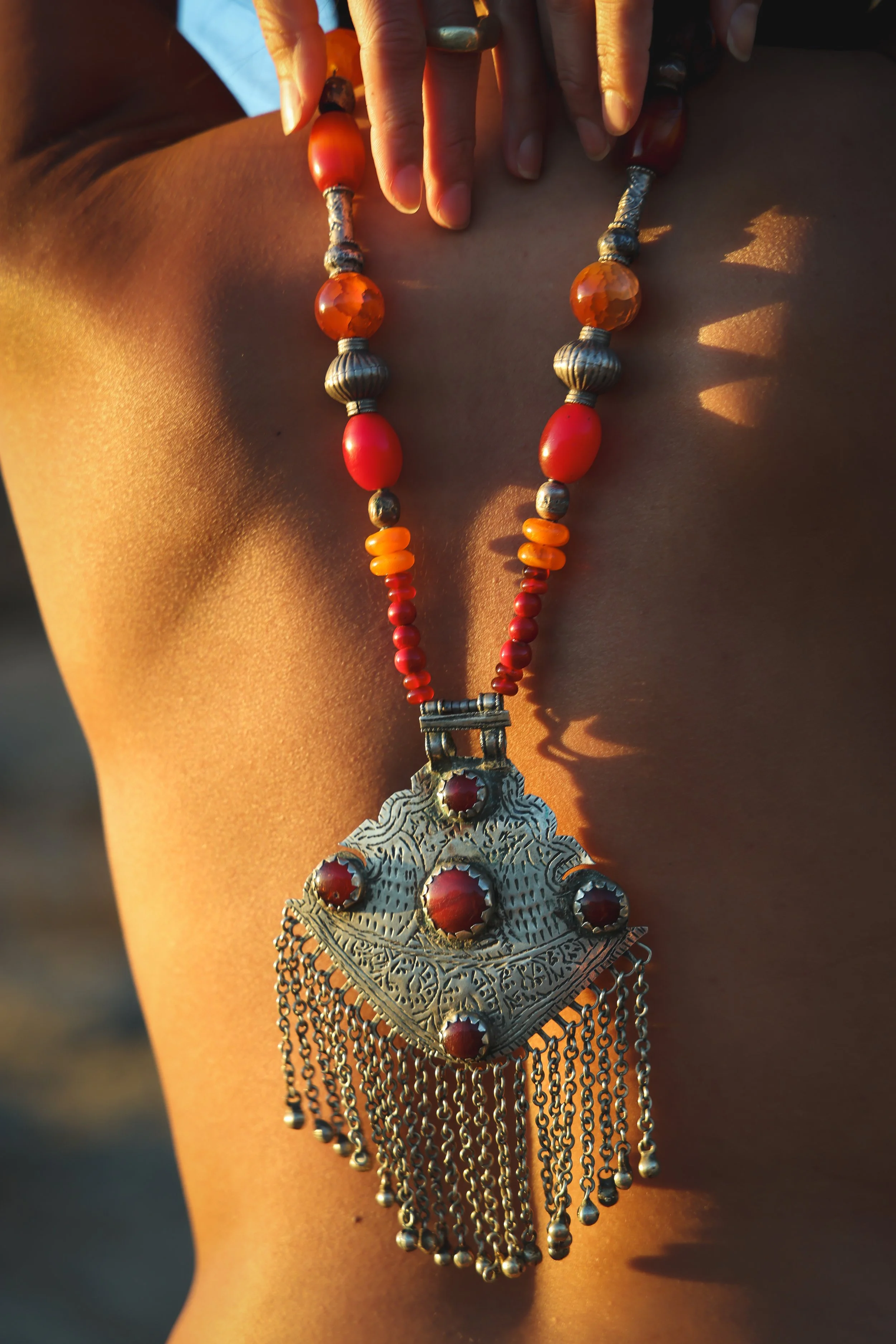 Close-up of a person wearing a large, ornate silver necklace with red gemstones, hanging chains, and intricate details on a person's shoulder and chest. The person's skin is warm-toned and sunlight highlights the jewelry.