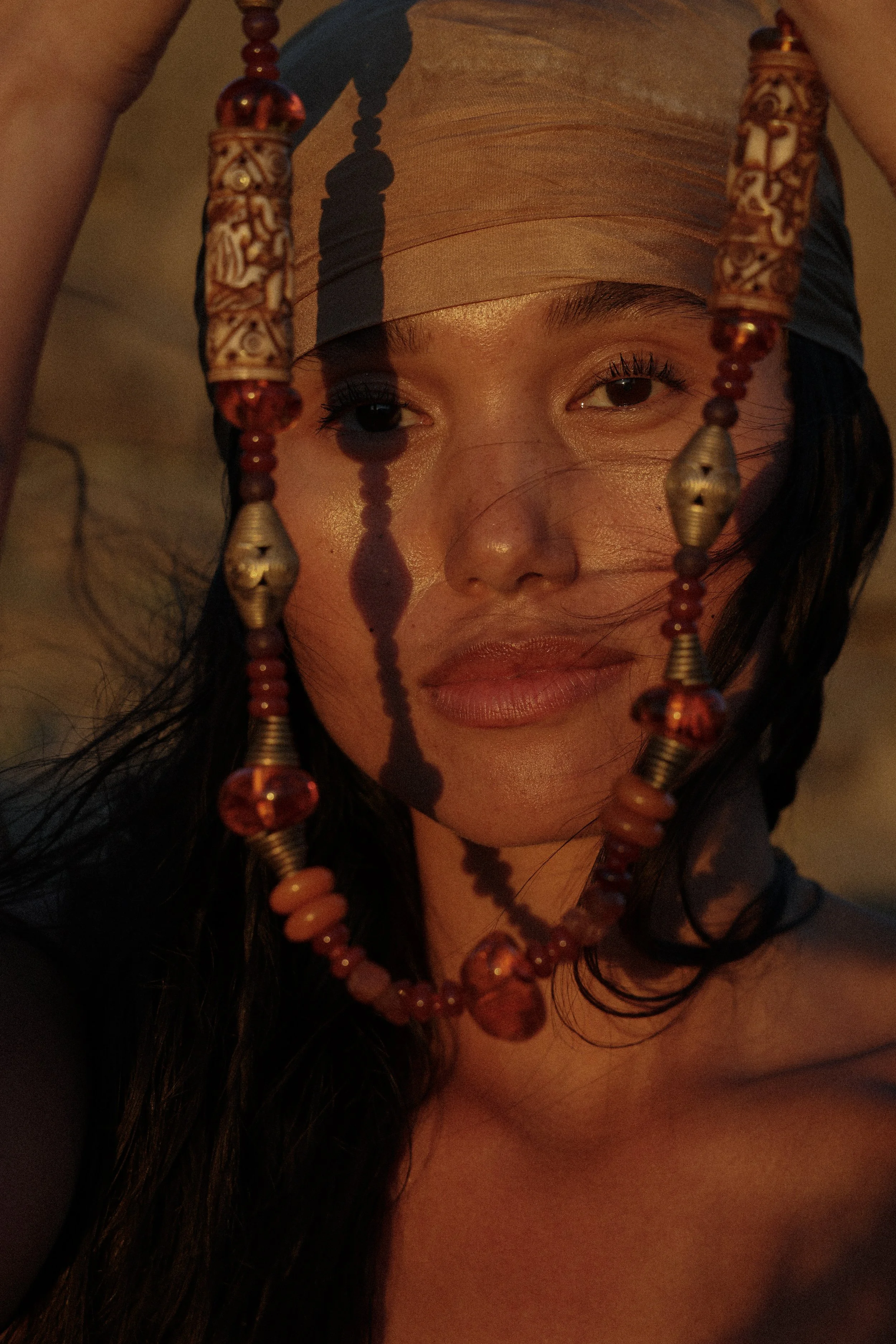 A woman wearing a beige headscarf and holding a beaded necklace with red, gold, and brown beads in front of her face, with part of her face and eye visible.