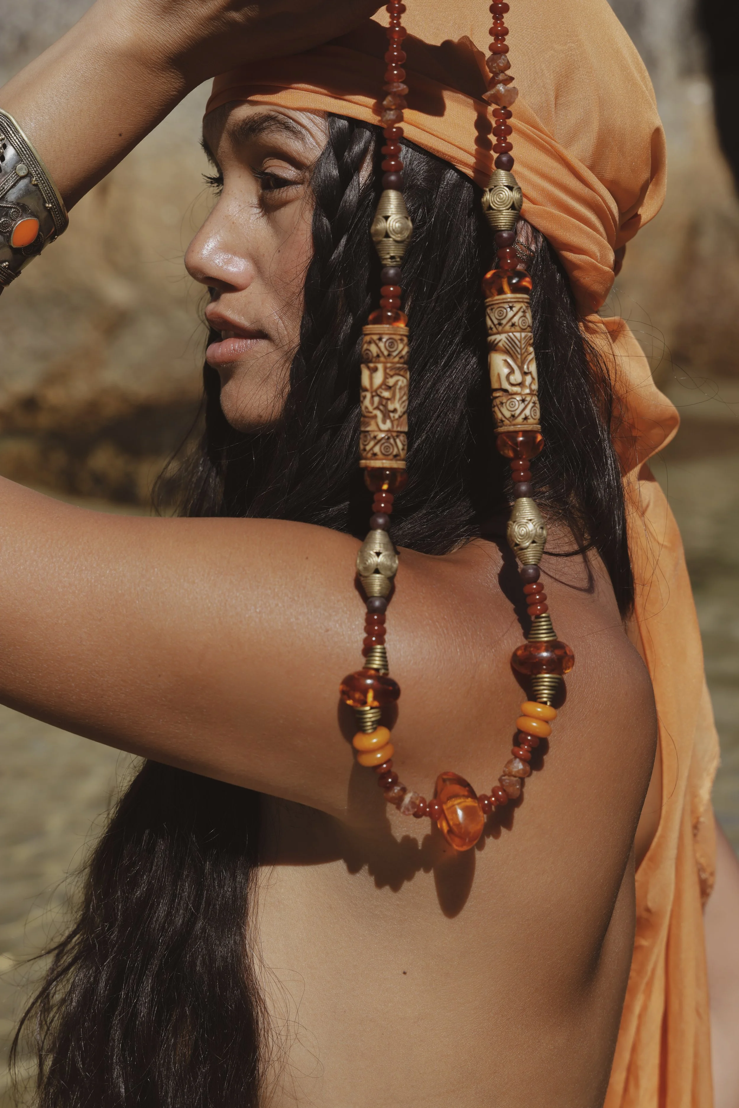 A woman with long dark hair wearing an orange headscarf and a beaded necklace, with her eyes closed and hand on her forehead, outdoors with a blurred natural background.