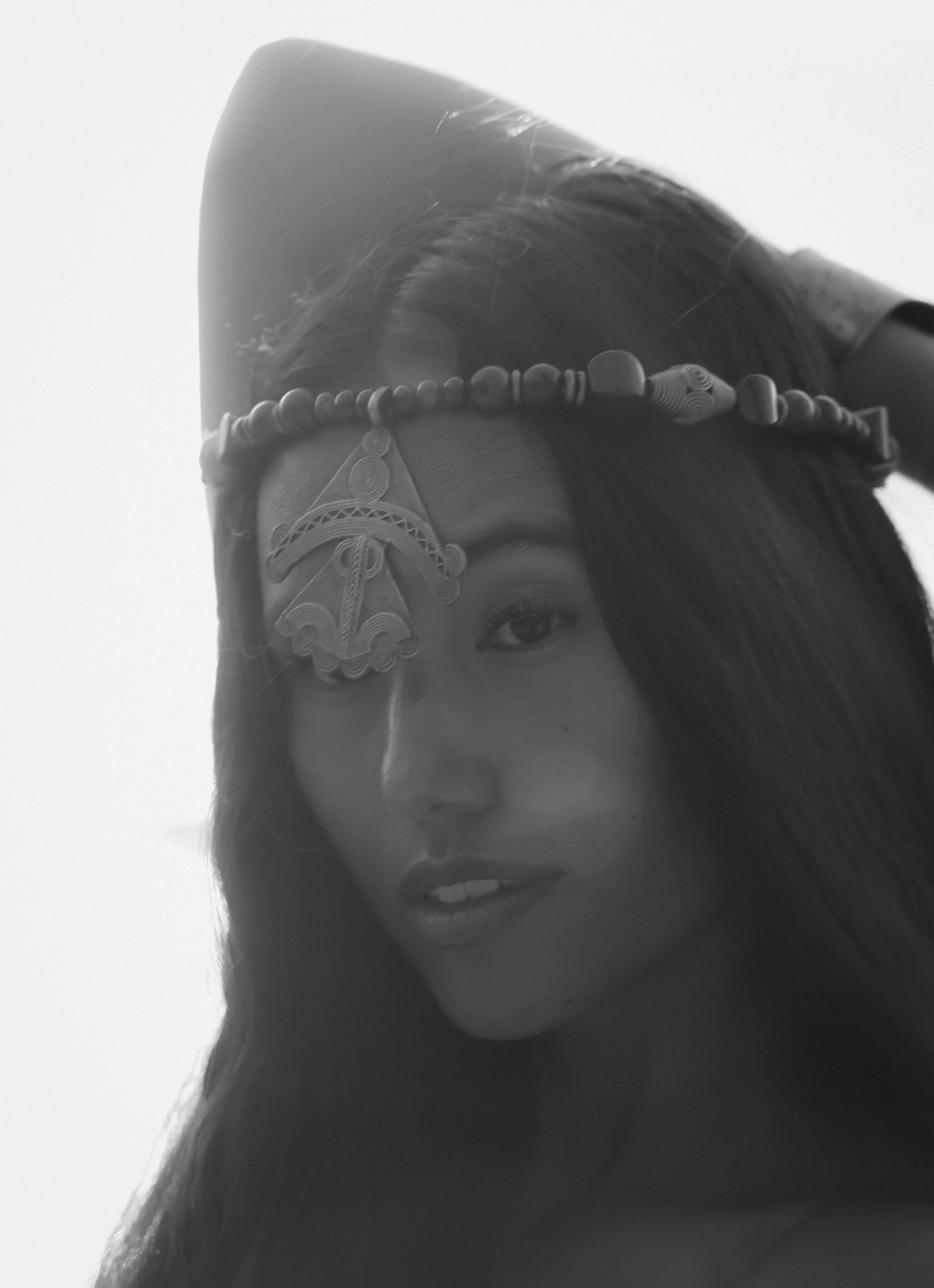 Close-up black and white photo of a woman's face partially covered with hair, wearing an ornate headband with beads and decorative elements.