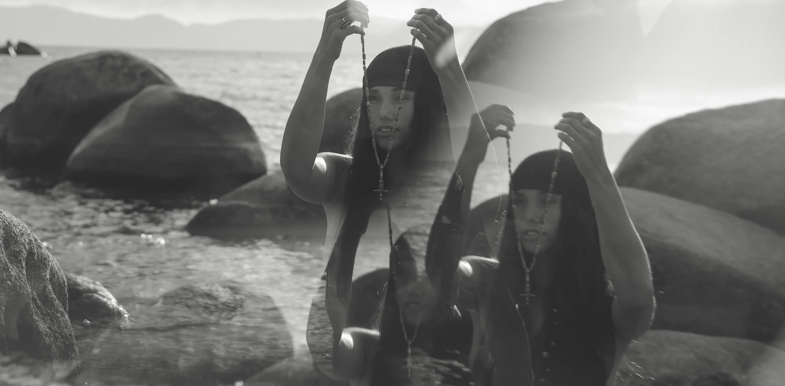Two women are standing in the water near rocks with the ocean in the background, holding beads and wearing dark clothing and headbands.