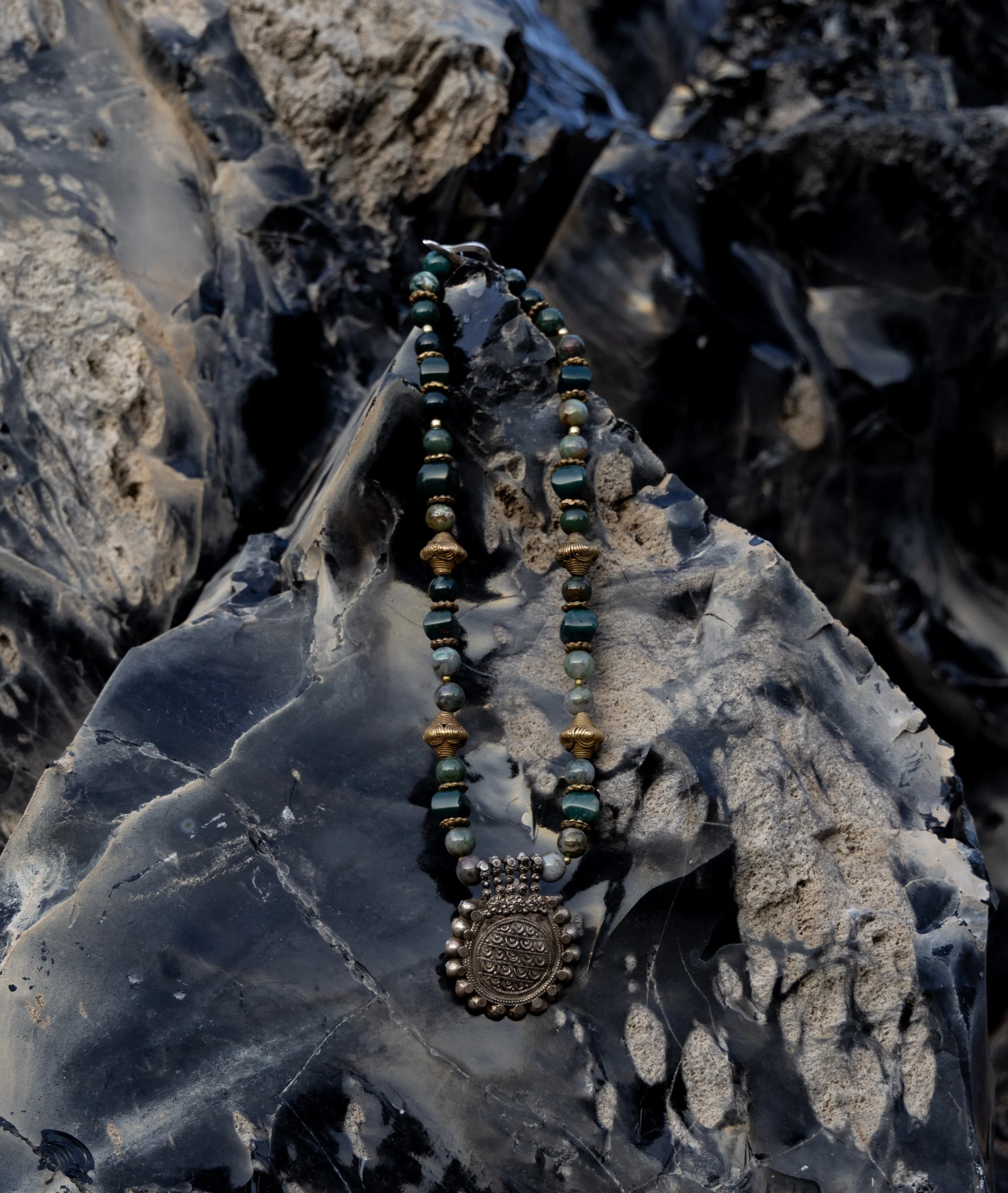 A piece of jewelry with green and white beads and a large ornate metal pendant resting on dark, textured rocks.
