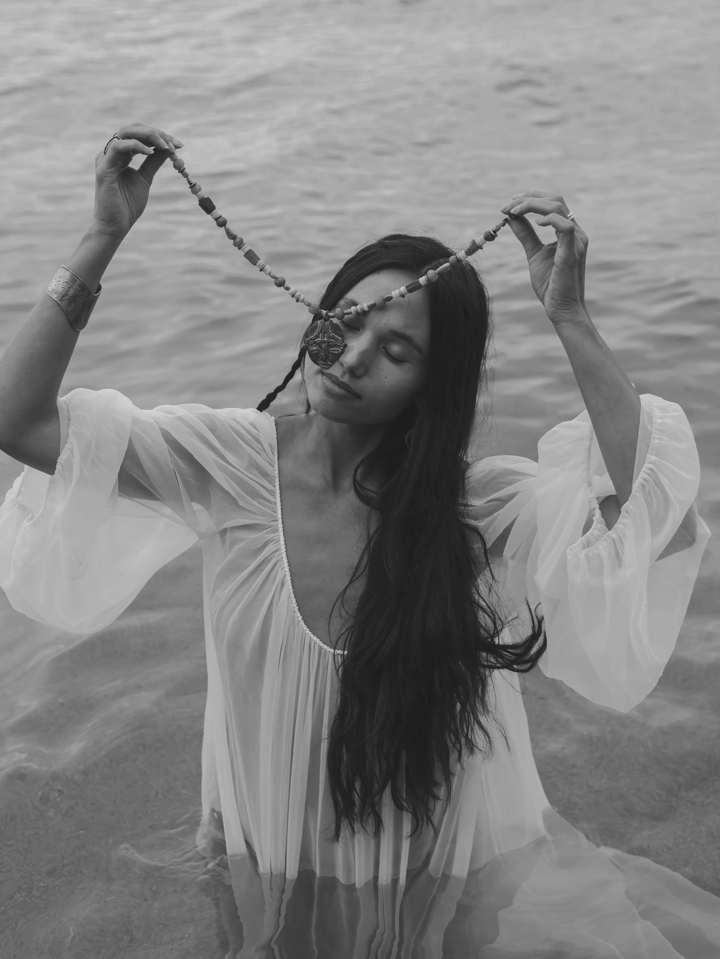 A woman with long dark hair standing in the water at the beach, holding a beaded necklace with a pendant over her face, with her eyes closed and wearing a loose, white, flowing dress.