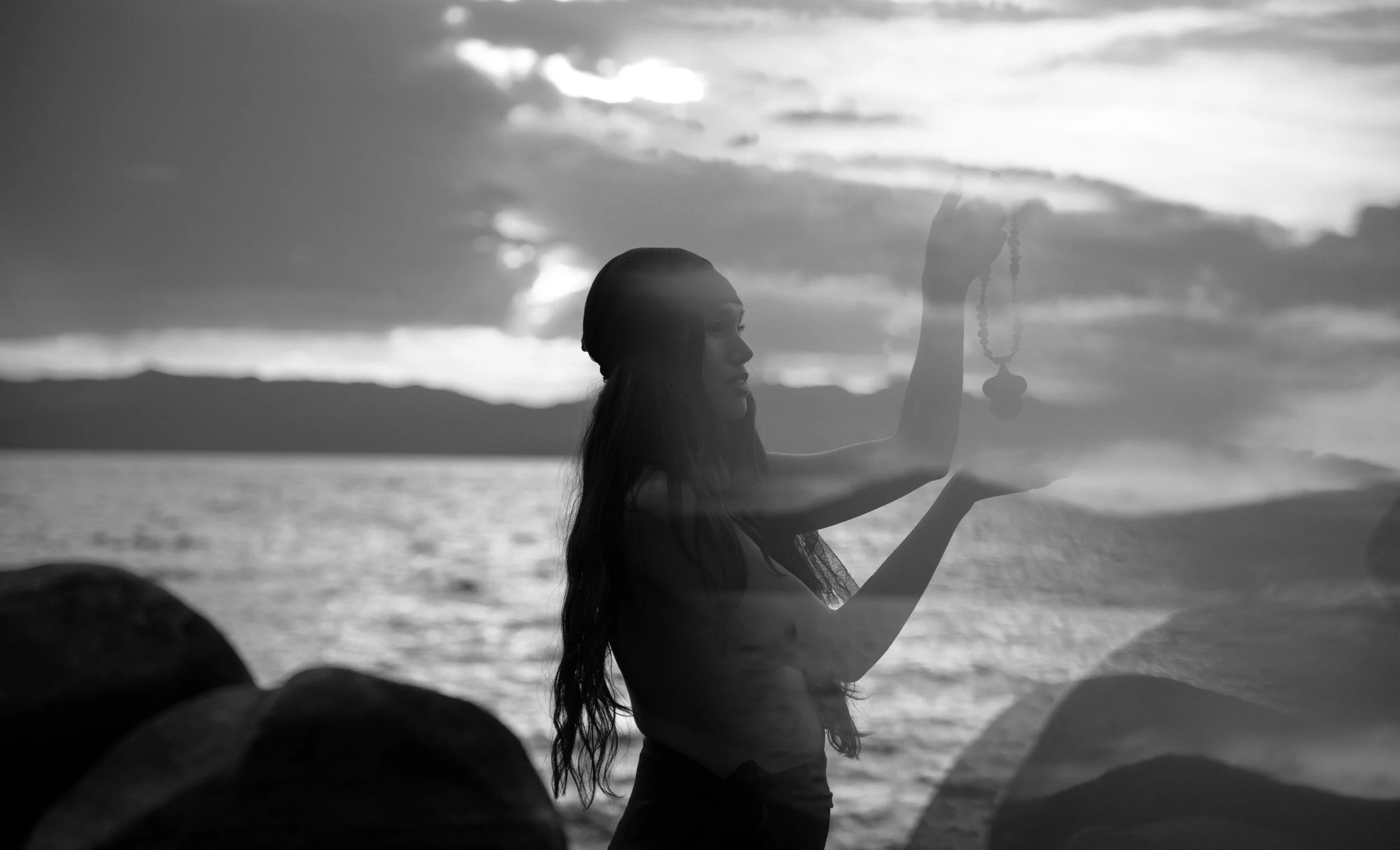 Black and white photo of a woman with long hair holding prayer beads in front of her face, standing on a beach at sunset with clouds in the sky.