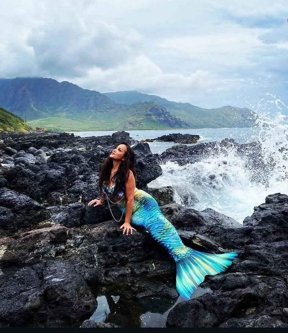 A woman with a mermaid tail resting on black volcanic rocks by the sea, with waves crashing and green mountains in the background under a cloudy sky.