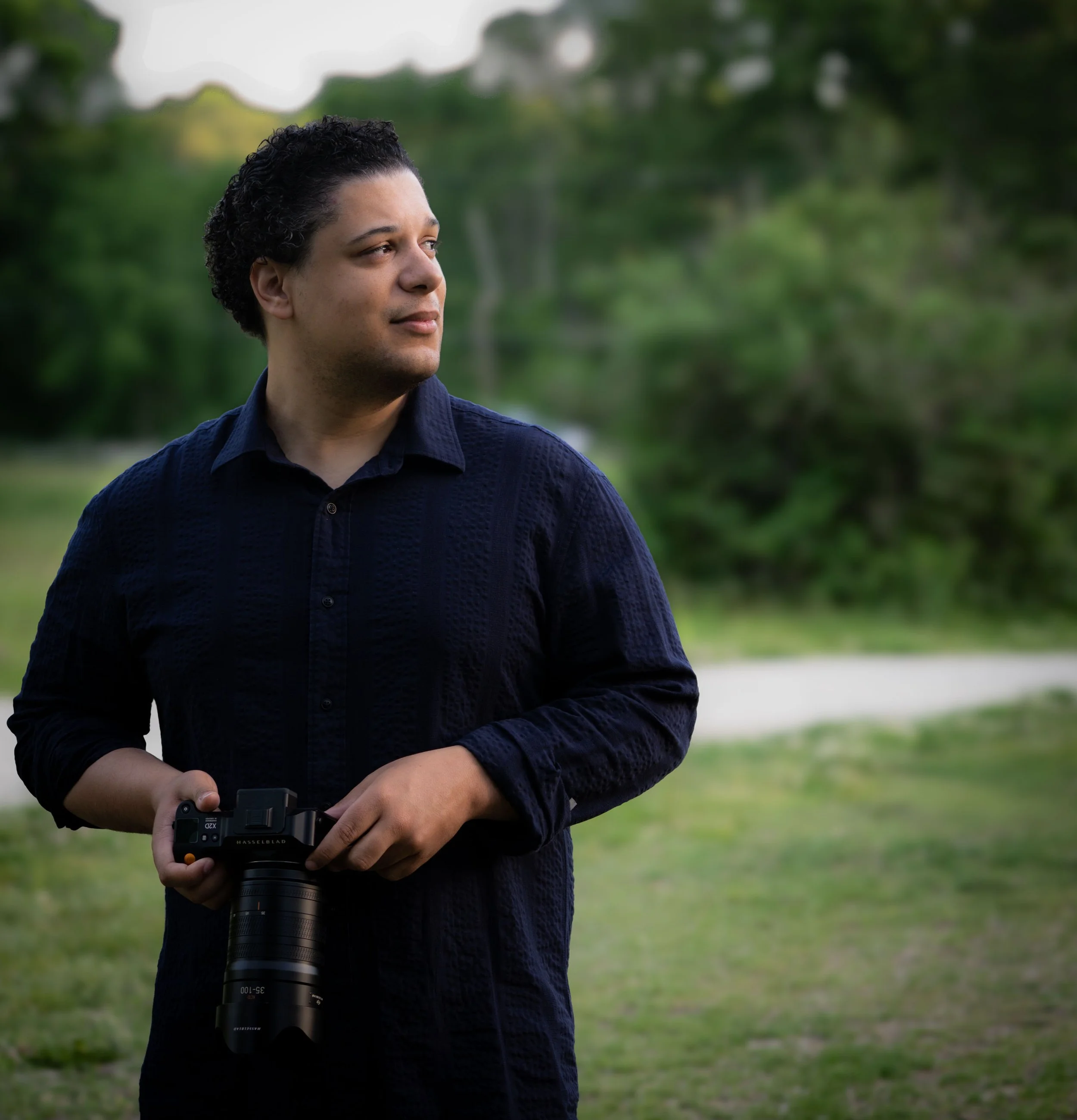 A man with dark, curly hair, wearing a dark blue shirt, standing outdoors with a blurry green natural background, holding a camera with a large telephoto lens.