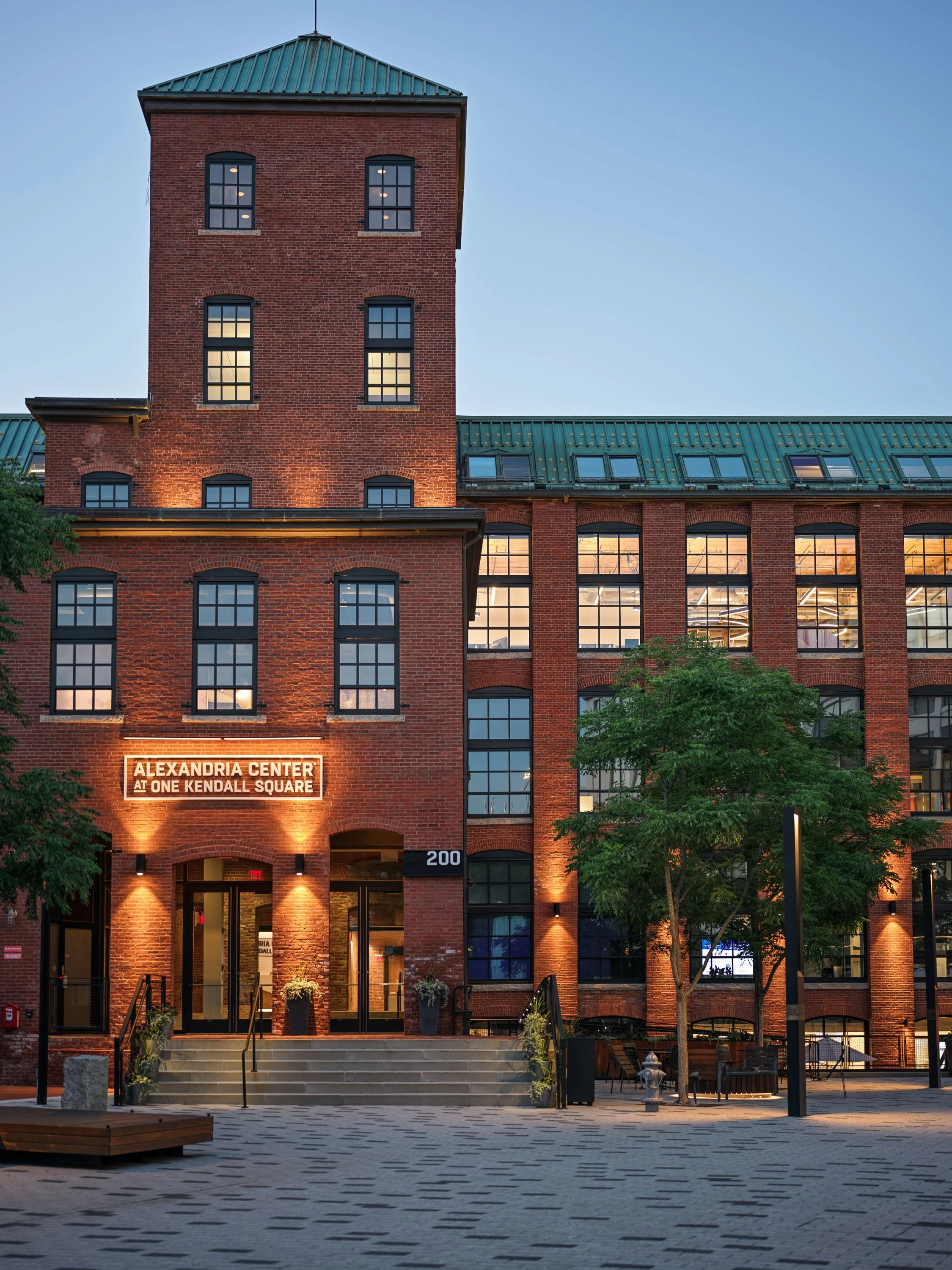 Exterior of a red brick building with large glass windows, illuminated with warm lighting, and a sign reading 'Alexandria Center at One Kendall Square' at dusk. There are trees, benches, and outdoor seating in the foreground.