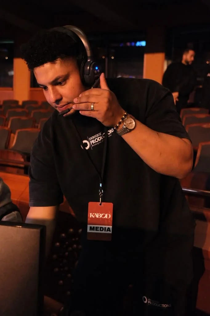 A man with curly black hair wearing a headset, a black T-shirt with a media badge, and a watch, is adjusting his headset in a dimly lit auditorium with empty seats.
