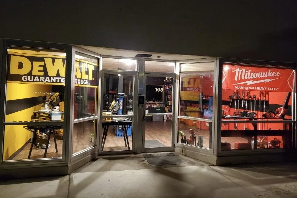 Exterior view of a store storefront at night with large colorful signs for DEWALT and Milwaukee power tools displayed in the windows.