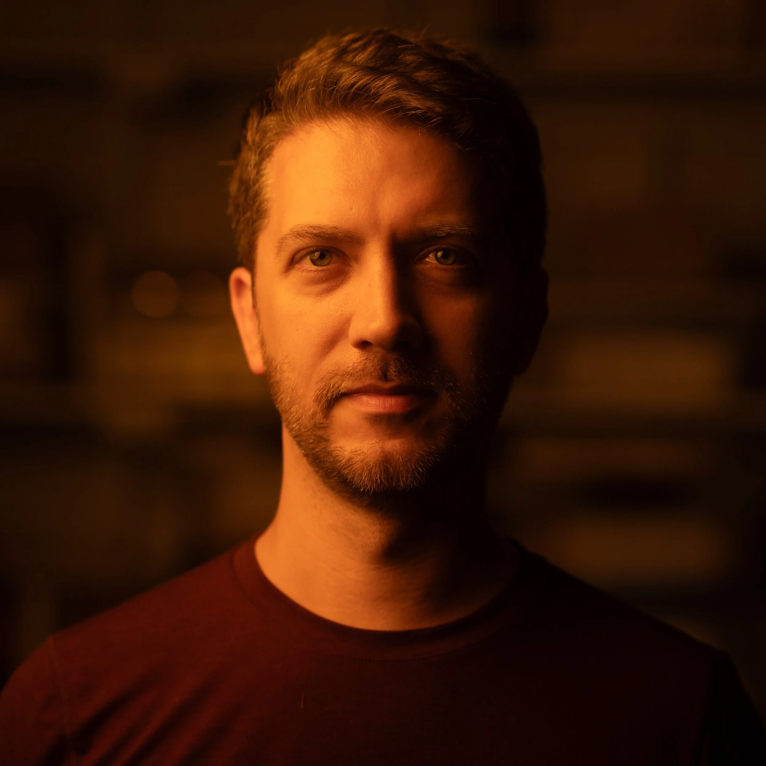 Close-up of Chris's face with short brown hair, green eyes, and a slight beard, illuminated by warm, orange lighting.