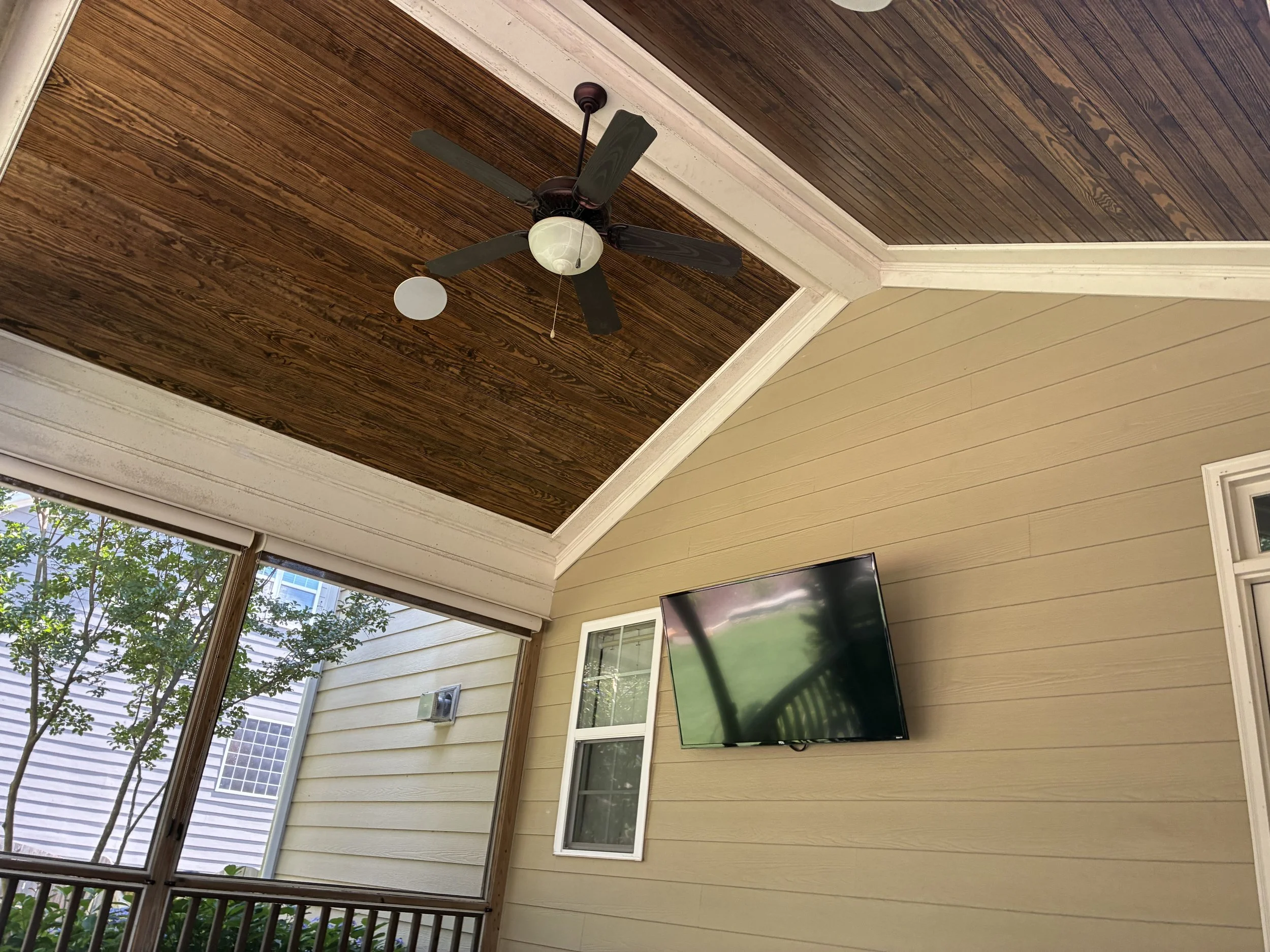 Outdoor patio with beige siding, large window with screen, mounted flat-screen TV, ceiling fan with dark blades, ceiling with wooden panels, and view of greenery outside.