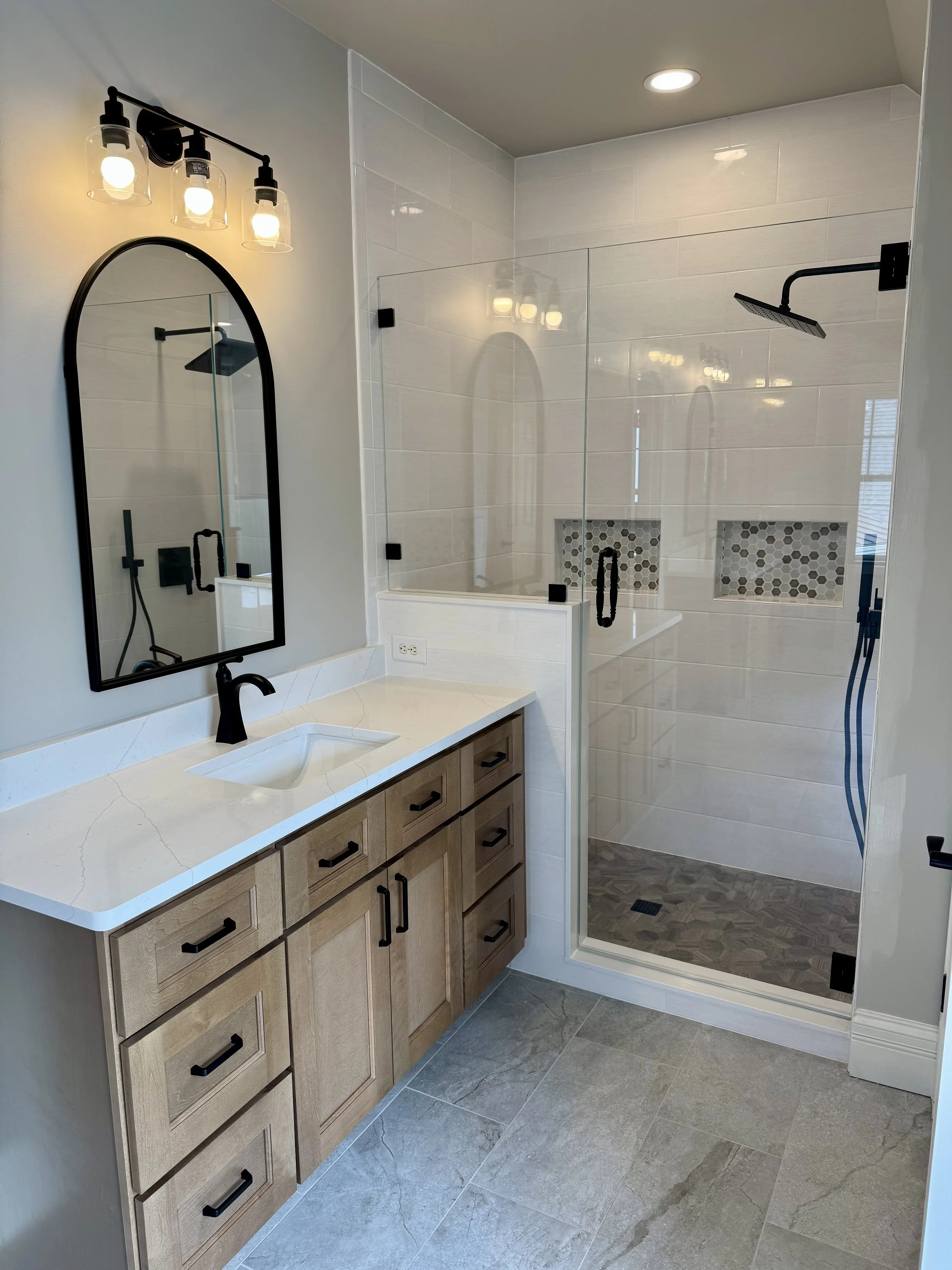 Wood bathroom vanity with a black faucet and arched mirror beside a glass shower.