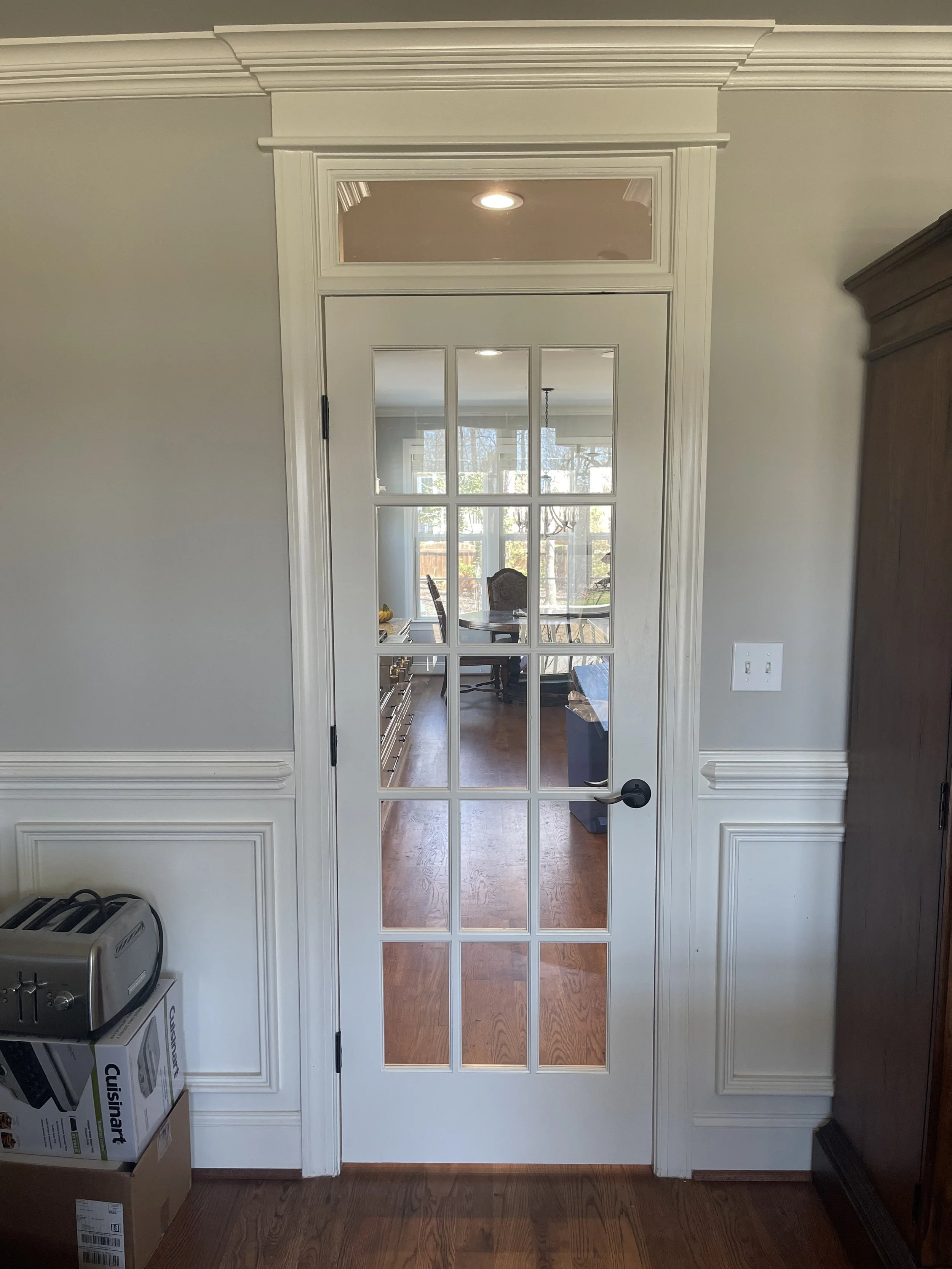 Interior view of a house through a glass-paneled door leading into a dining room with hardwood floors and windows showing trees outside.