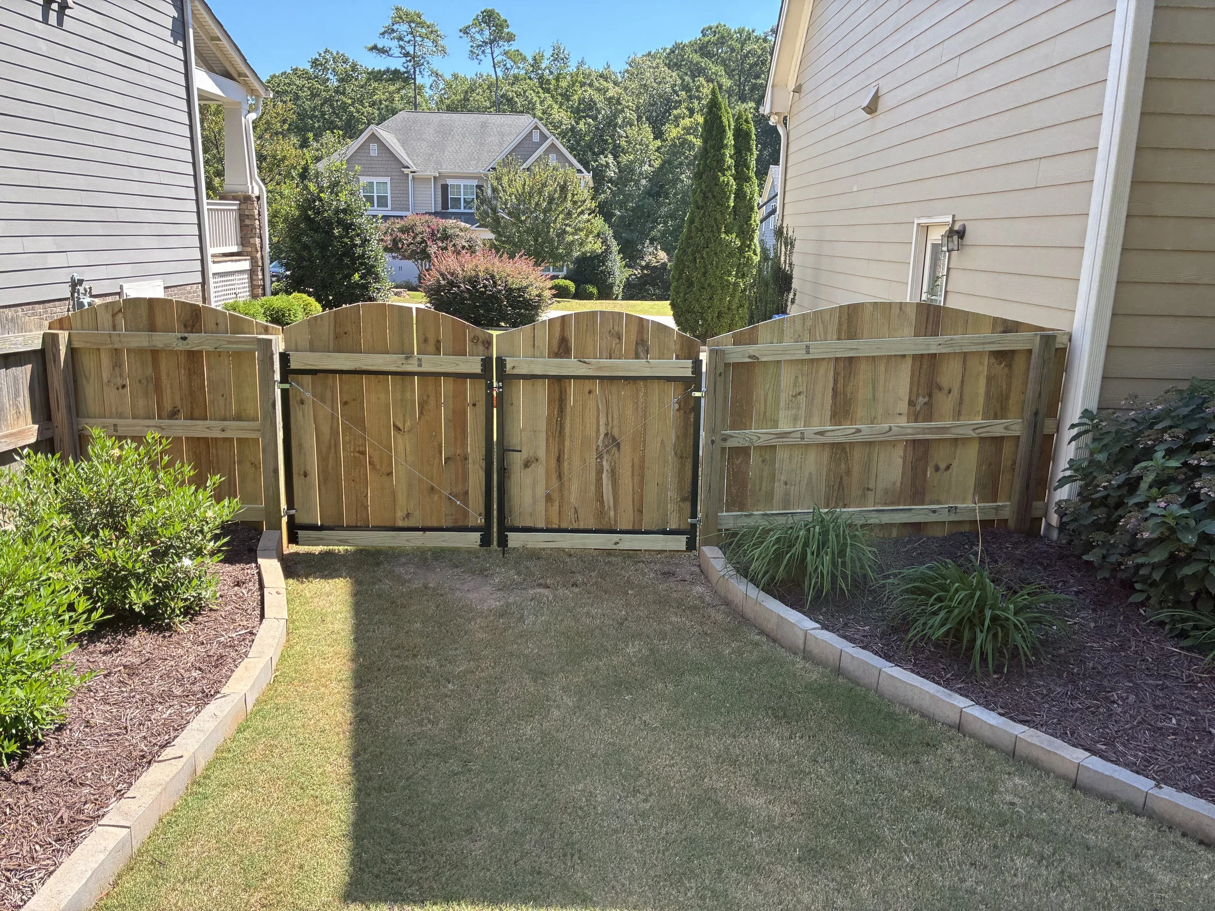 View of a backyard with a wooden fence, a well-maintained lawn, and flower beds on either side, with houses and trees in the background under a clear blue sky.