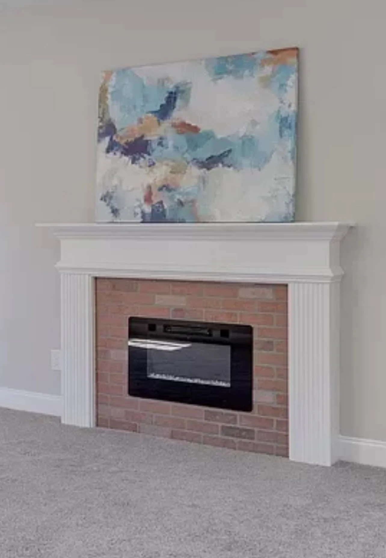 Living room fireplace with a white mantel, red brick surround, black insert, and a large abstract painting with blue, white, and beige colors hanging above it, with a carpeted floor.