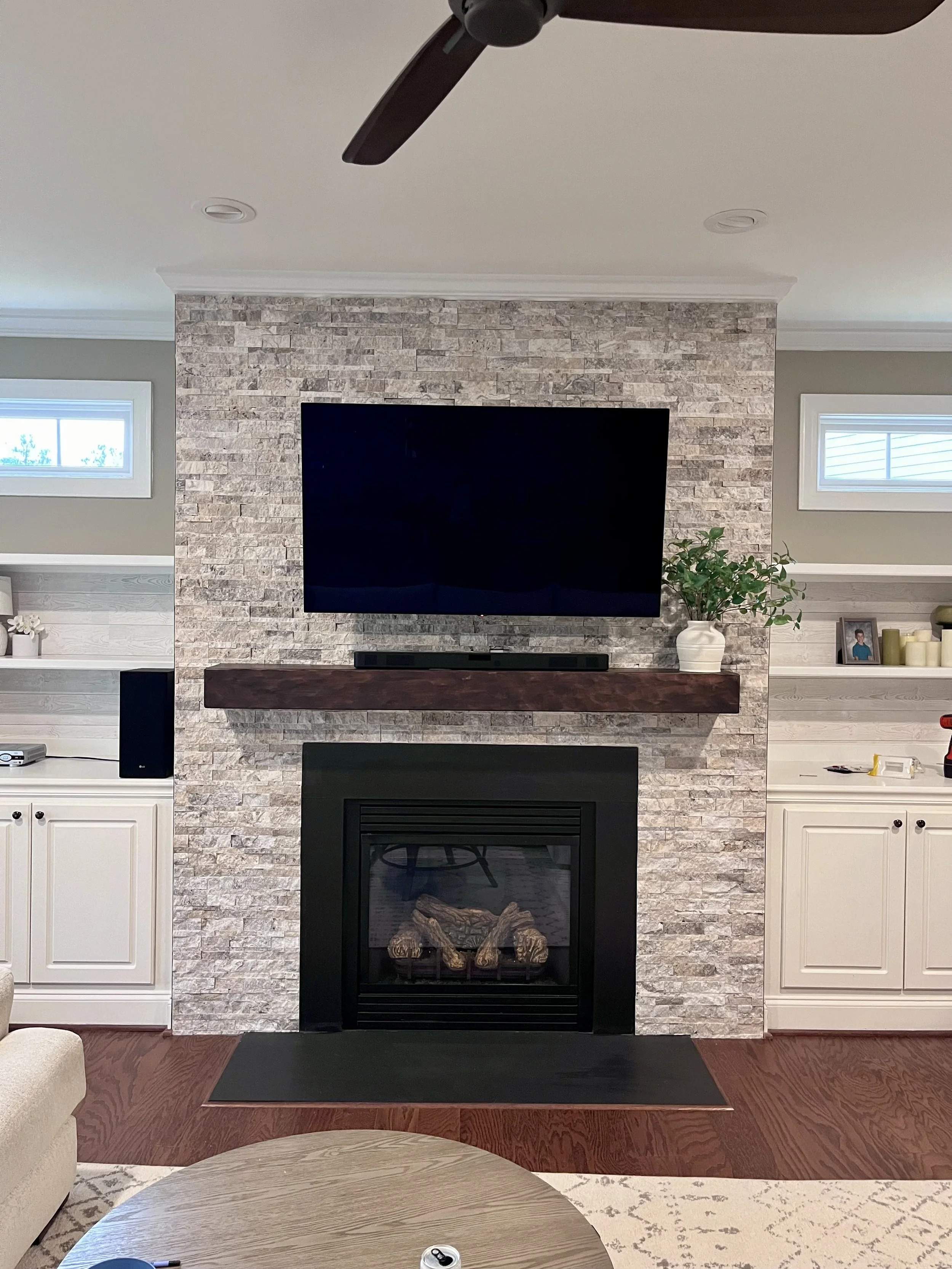 Living room with stone fireplace, mounted TV, mantel shelf, potted plant, framed photo, and decorative candles, seen from an angle with a coffee table and part of an armchair in the foreground.