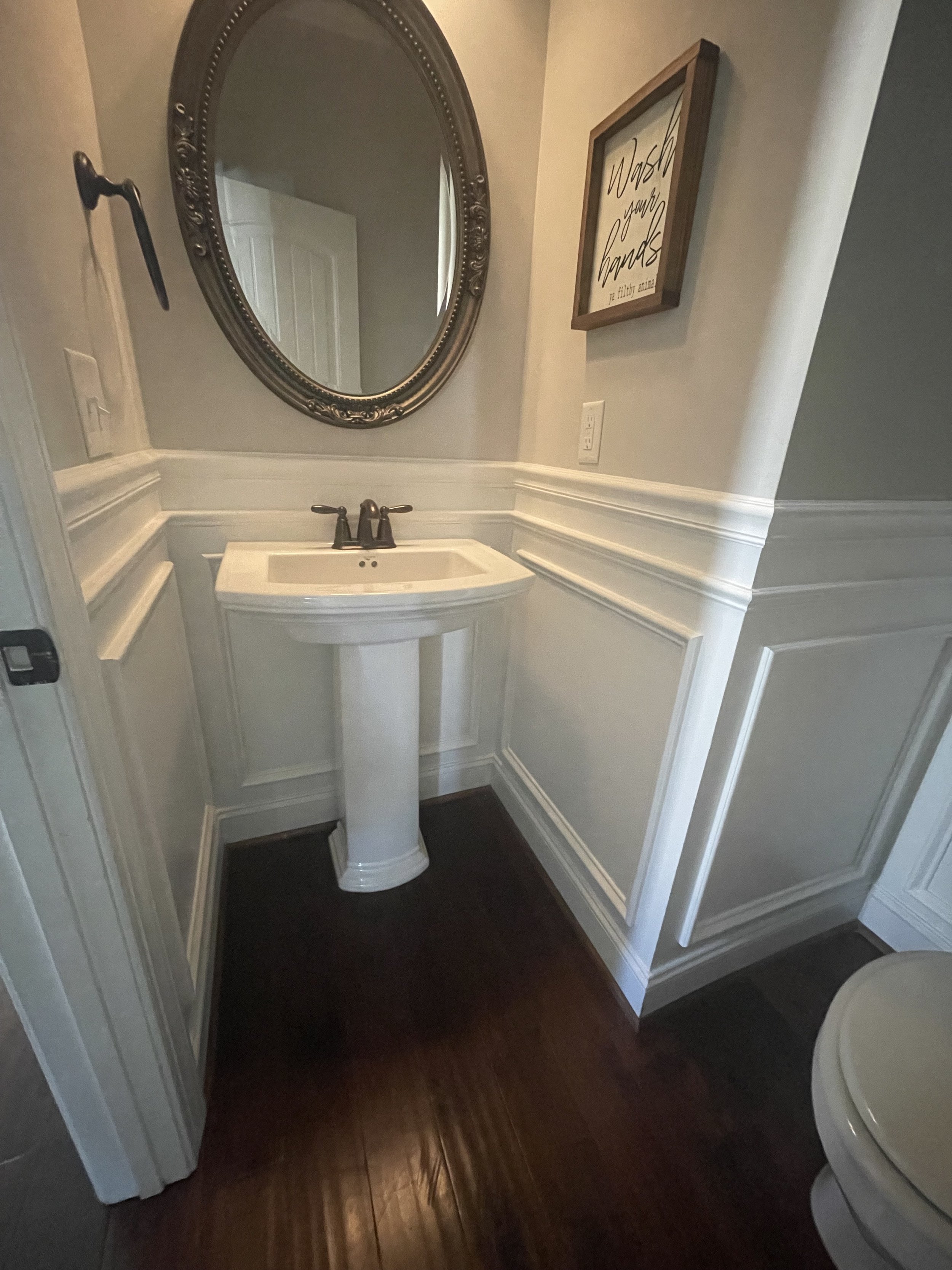 A small bathroom with a white pedestal sink, a large oval mirror with an ornate frame, a framed wall art saying 'Dreams for wash', and wood flooring.