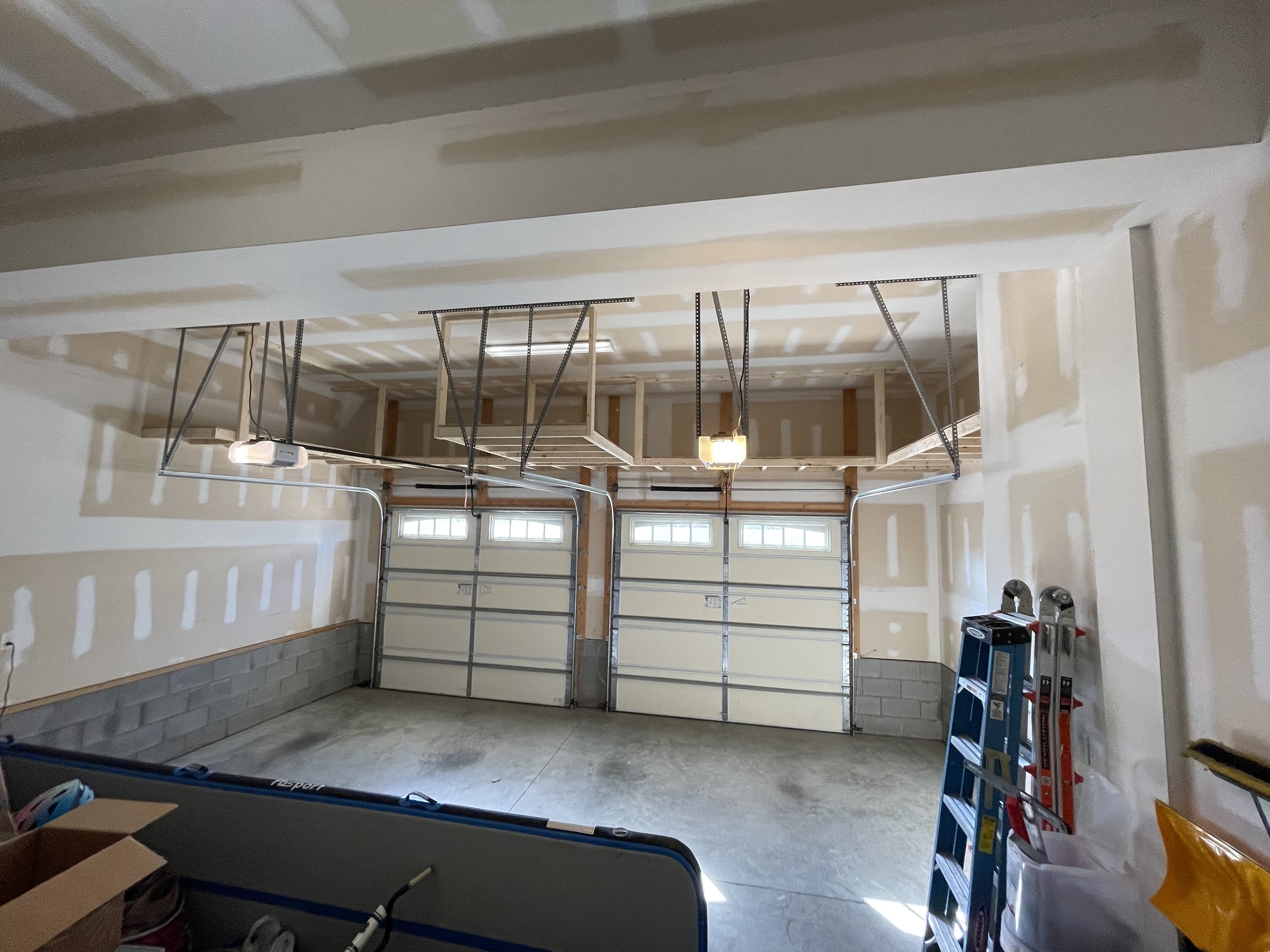 Garage with unfinished drywall walls, an overhead garage door, construction tools, and a blue ladder.