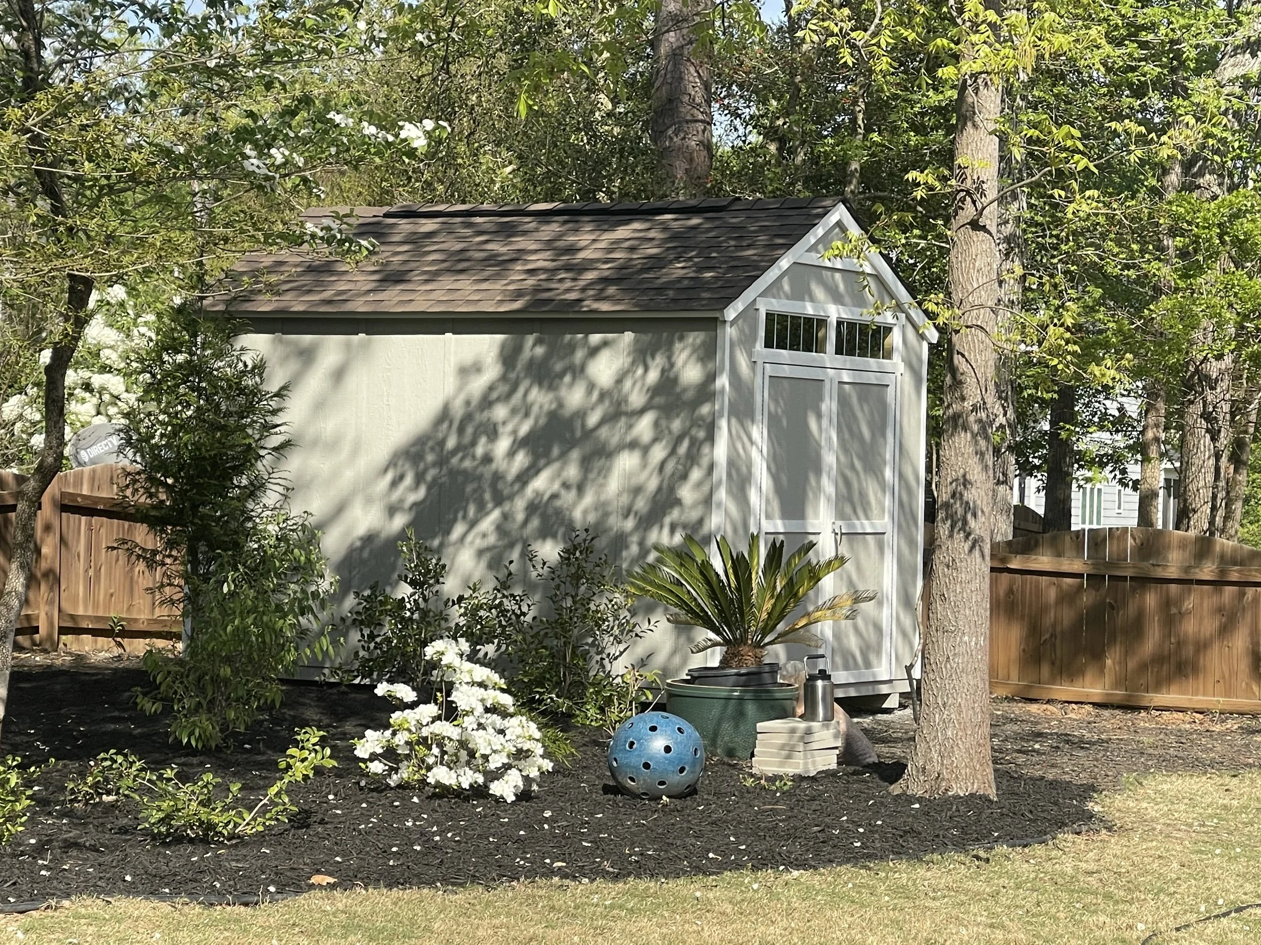 A backyard with a white shed, surrounded by trees and plants, including bushes with white flowers. There are gardening items on the ground such as a blue ball, a green planter, and other containers. A wooden fence runs along the back of the yard.