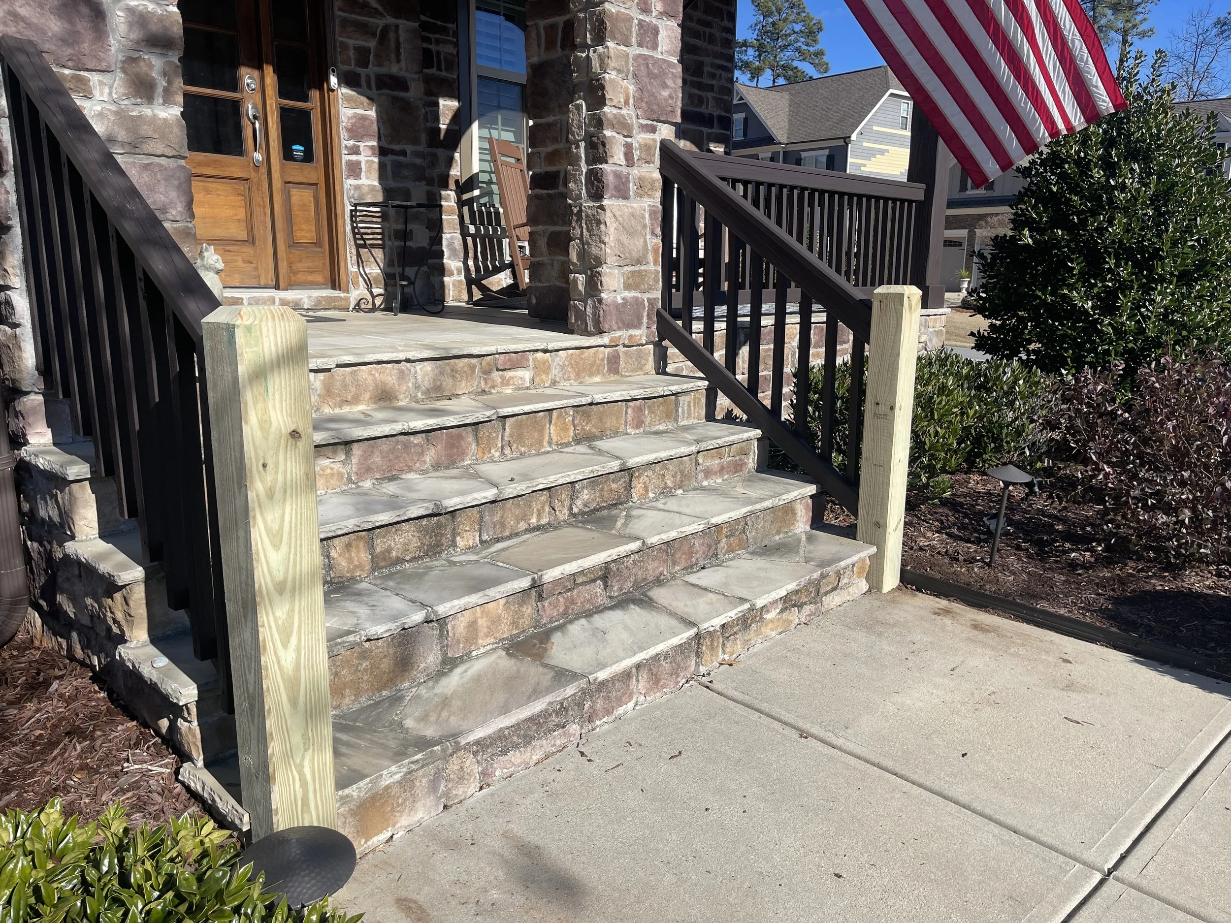 Front porch with stone steps leading to a wooden door, a rocking chair, an American flag, and greenery.