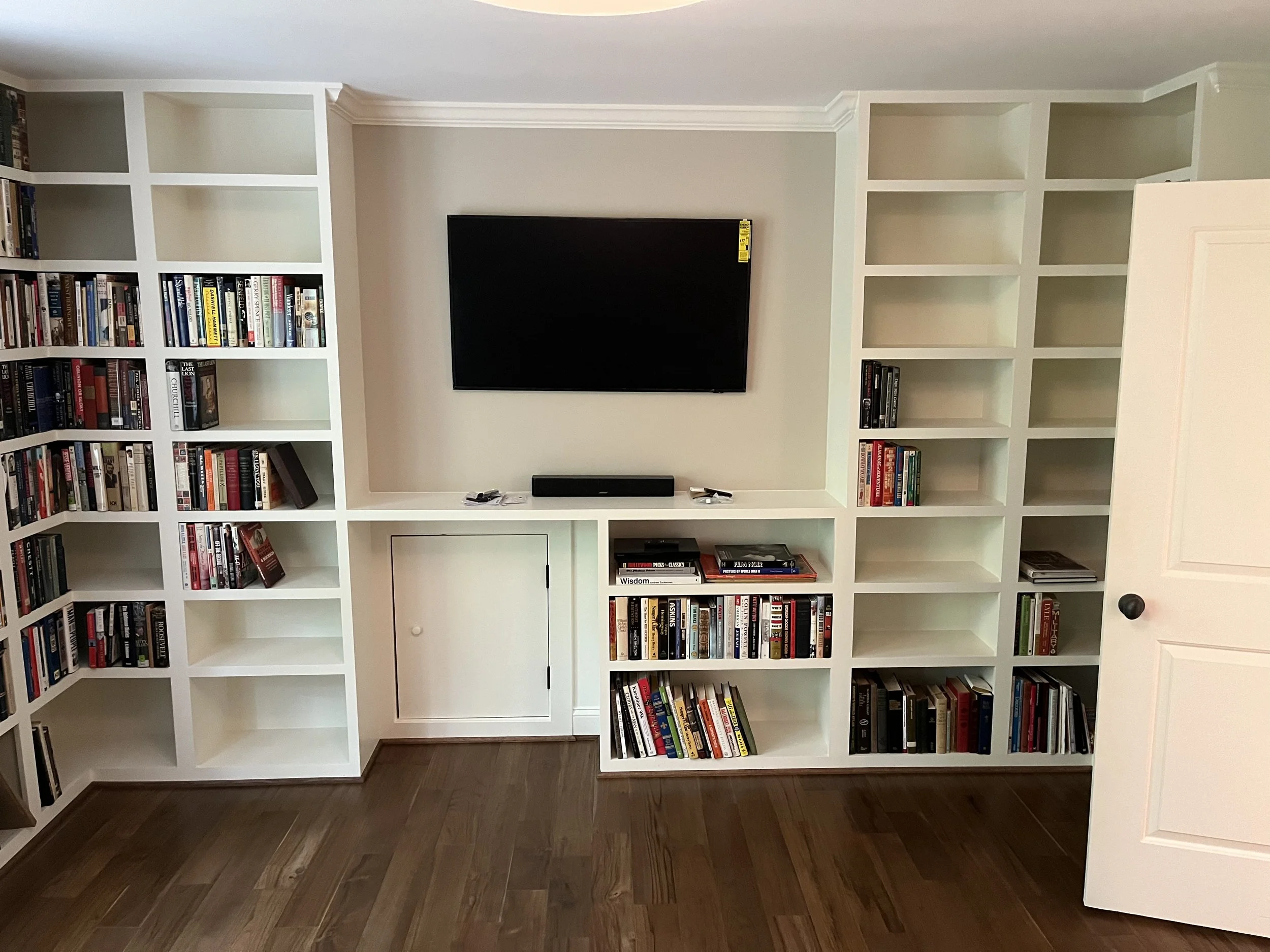 A white bookshelf with multiple compartments filled with books. A flat-screen TV is mounted in the center of the bookshelf on a beige wall. The room has dark hardwood floors.