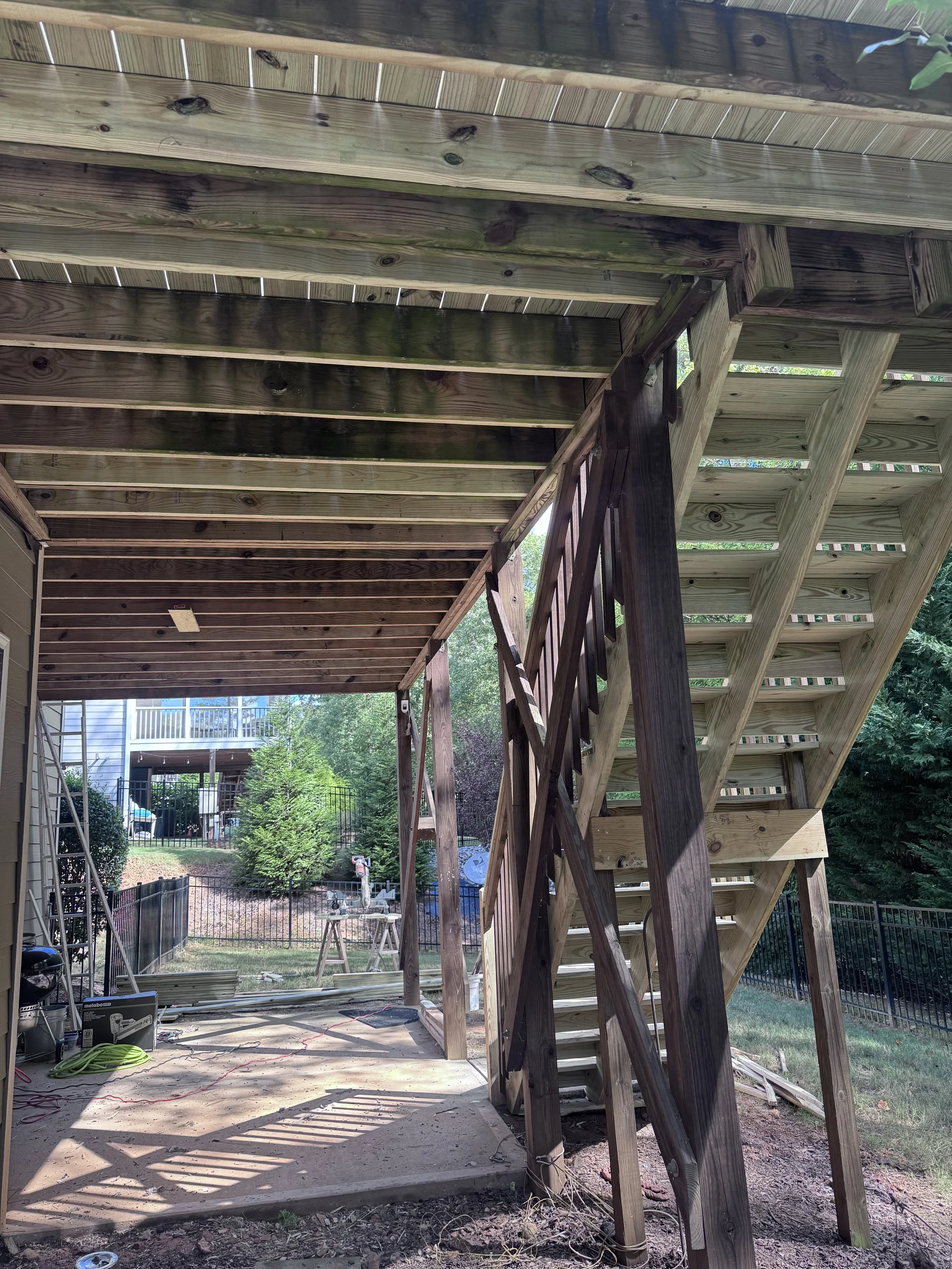 Under construction backyard deck with unfinished staircase and support beams, sunlit outdoors, construction tools, and materials scattered around.
