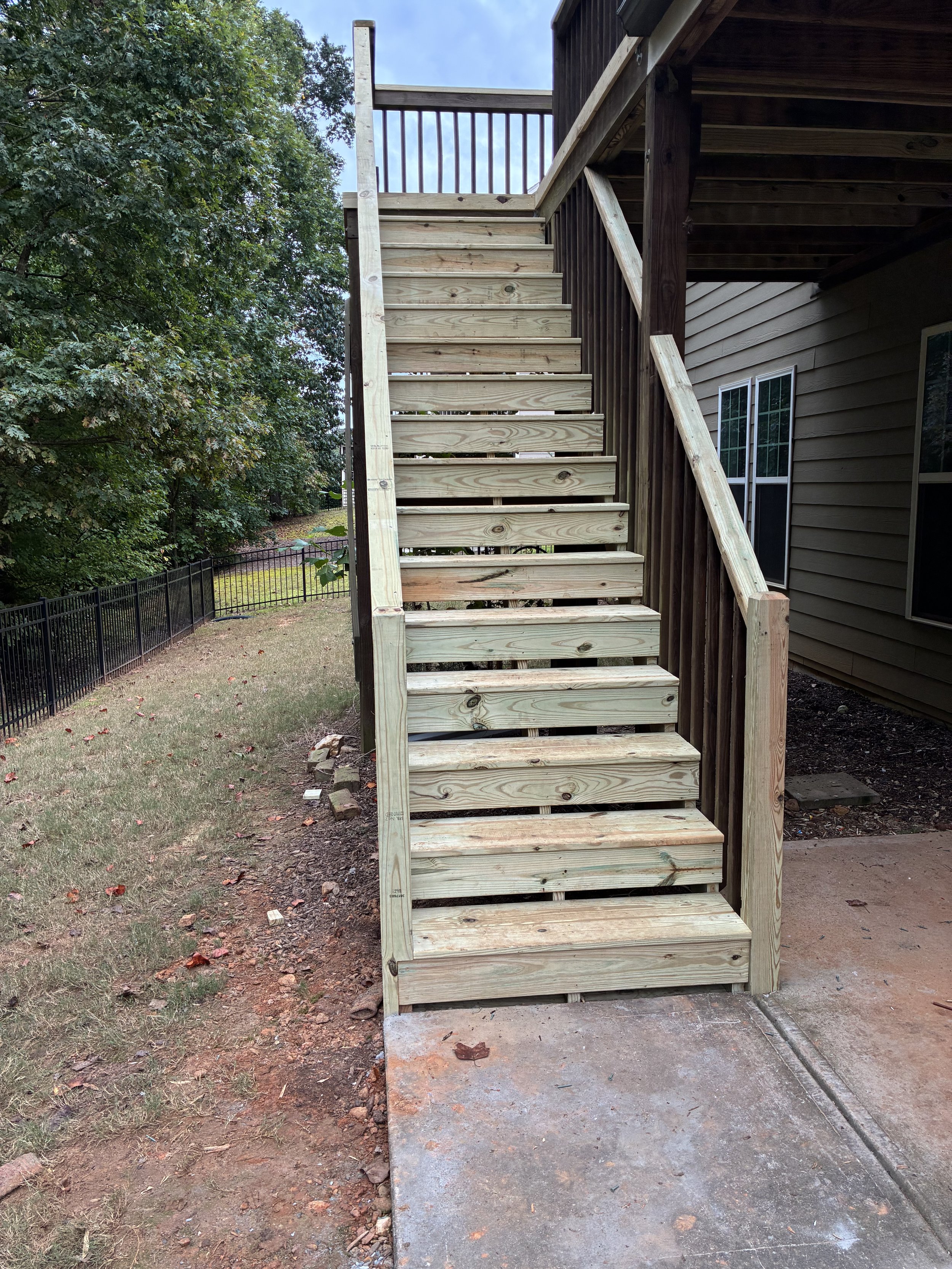 A new wooden staircase attached to the back of a house, leading up to a second floor balcony with a wooden railing. The staircase is outdoors, with trees and a black fence in the background, and a mix of grass and dirt on the ground.