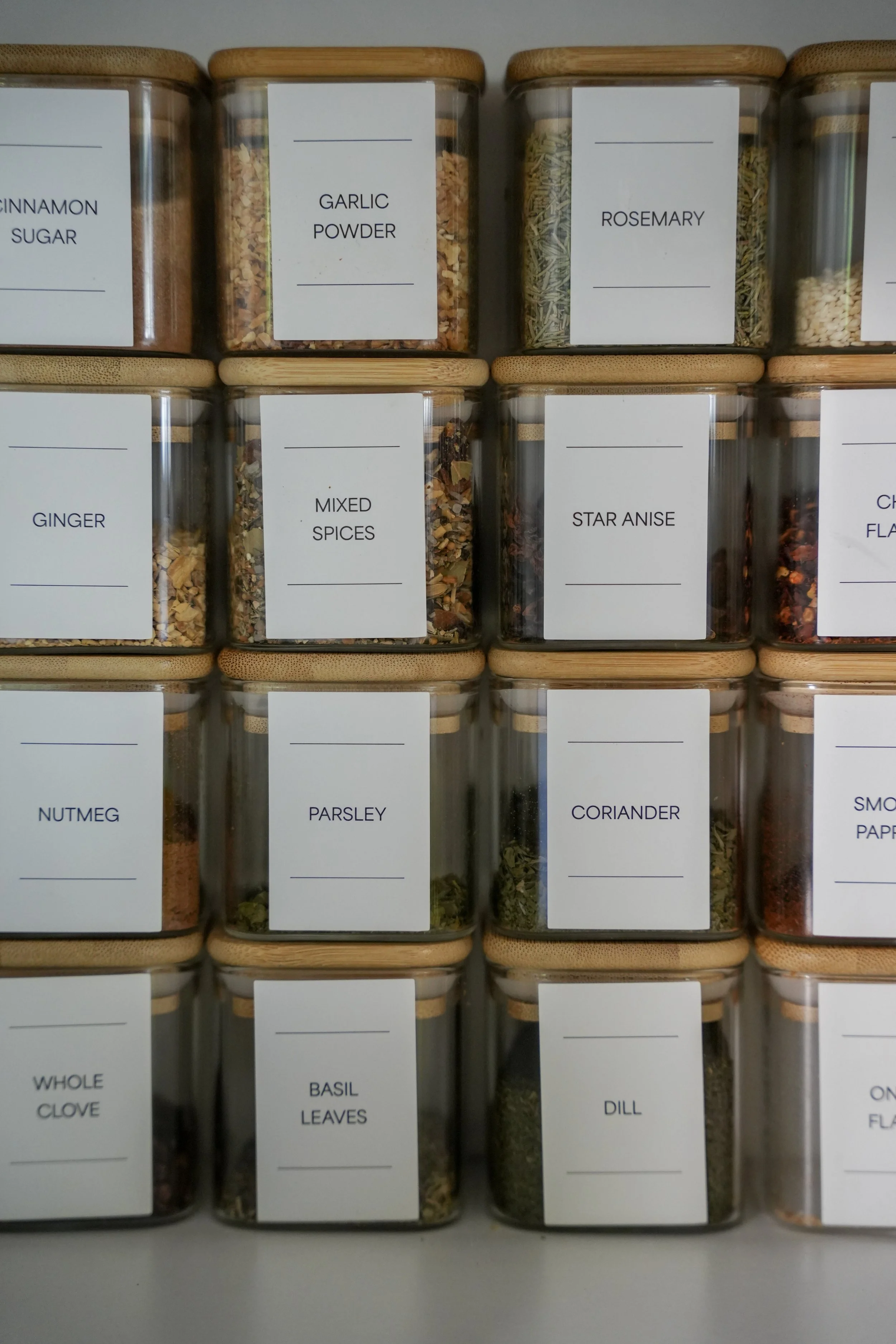 Various spices stored in glass jars with beige bamboo lids, labeled with their names including garlic powder, rosemary, mixed spices, star anise, ginger, parsley, coriander, whole clove, basil leaves, dill, and smoked paprika.