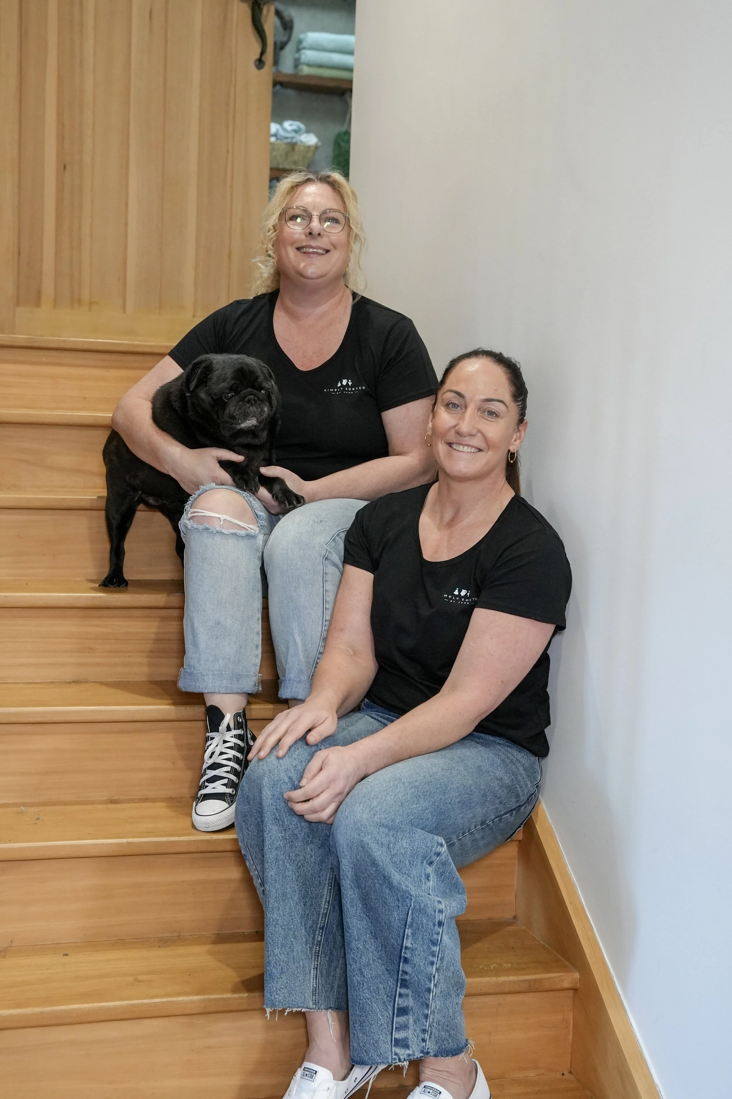 Two women sitting on wooden stairs with a small black French Bulldog. Both women are smiling and wearing black shirts and jeans. One woman has blonde hair and glasses, and the other has dark hair tied back. The background features a plain white wall and a wooden shelf with towels.