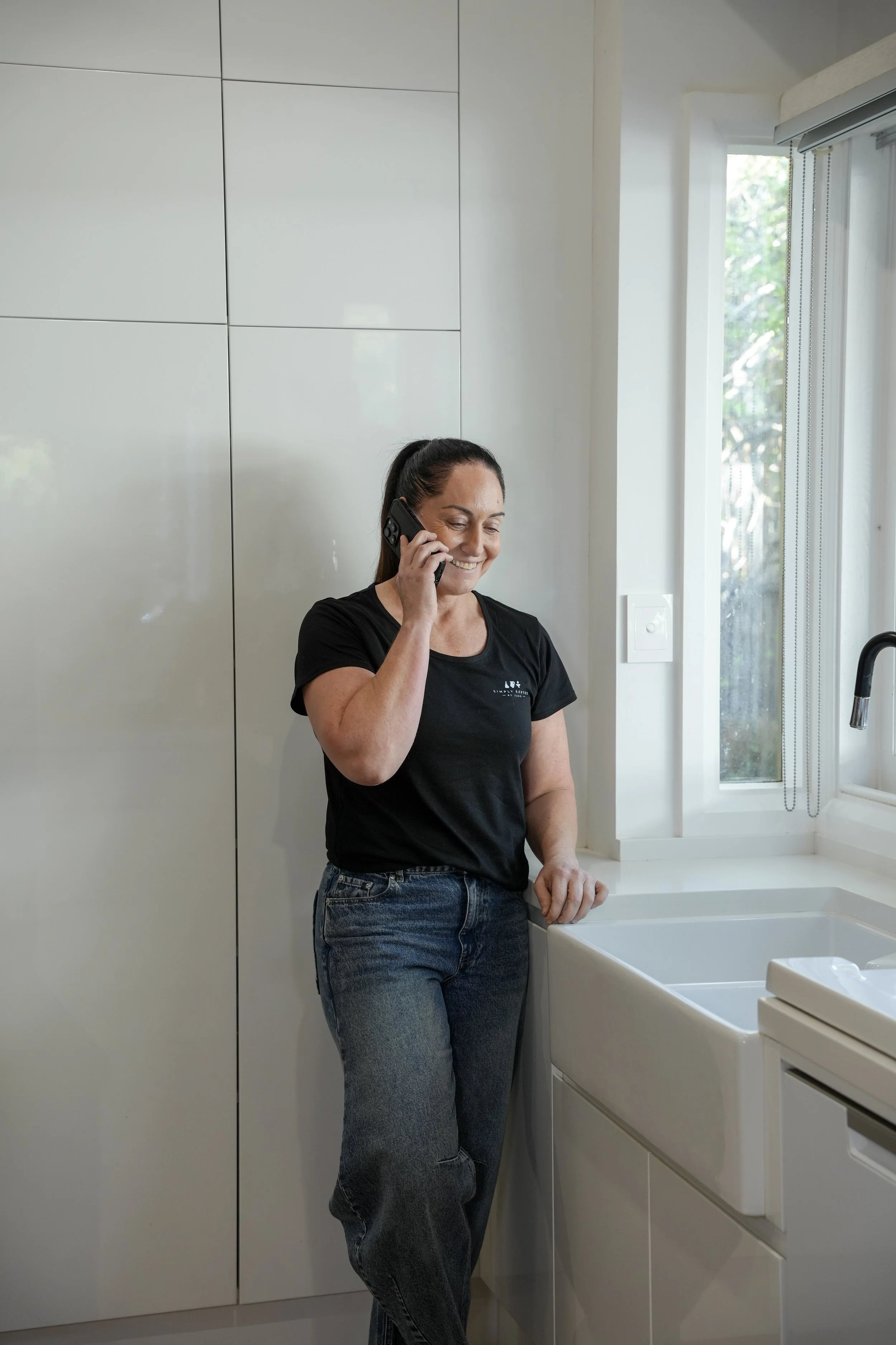 A woman with a ponytail talking on a cell phone in a bright kitchen, dressed in a black t-shirt and blue jeans, standing next to a white sink and white cabinetry near a window.