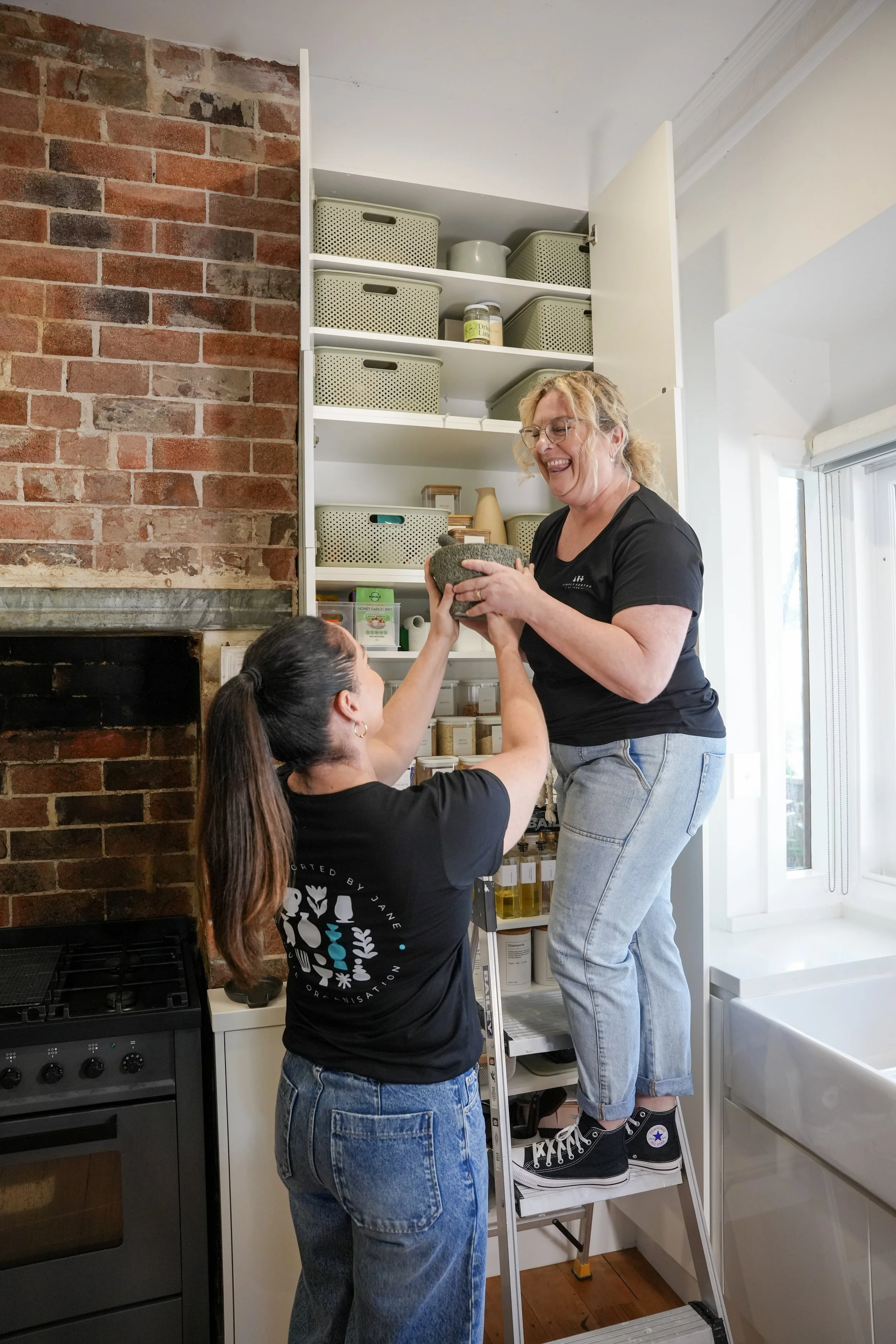 Two women are in a kitchen, one is standing on a step ladder and the other is holding her hand. The woman on the ladder is smiling and wearing black Converse sneakers. The woman on the ground has long brown hair and is wearing a black t-shirt and blue jeans. The kitchen has a brick wall, open shelving with storage baskets, and a stove.