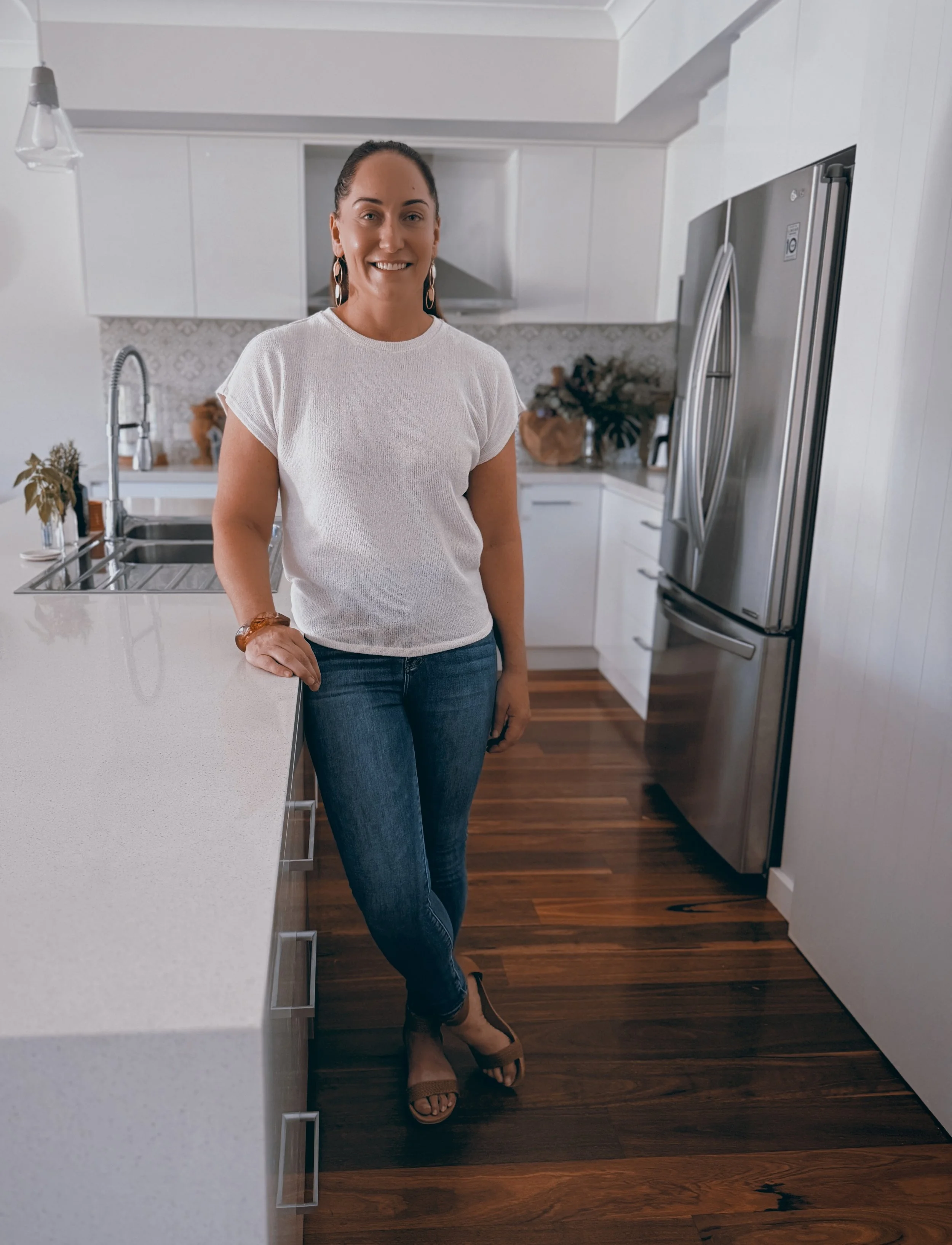 A woman with dark hair and earrings wearing a white top and blue jeans, standing in a modern kitchen with white cabinets, a stainless steel refrigerator, and hardwood floors.