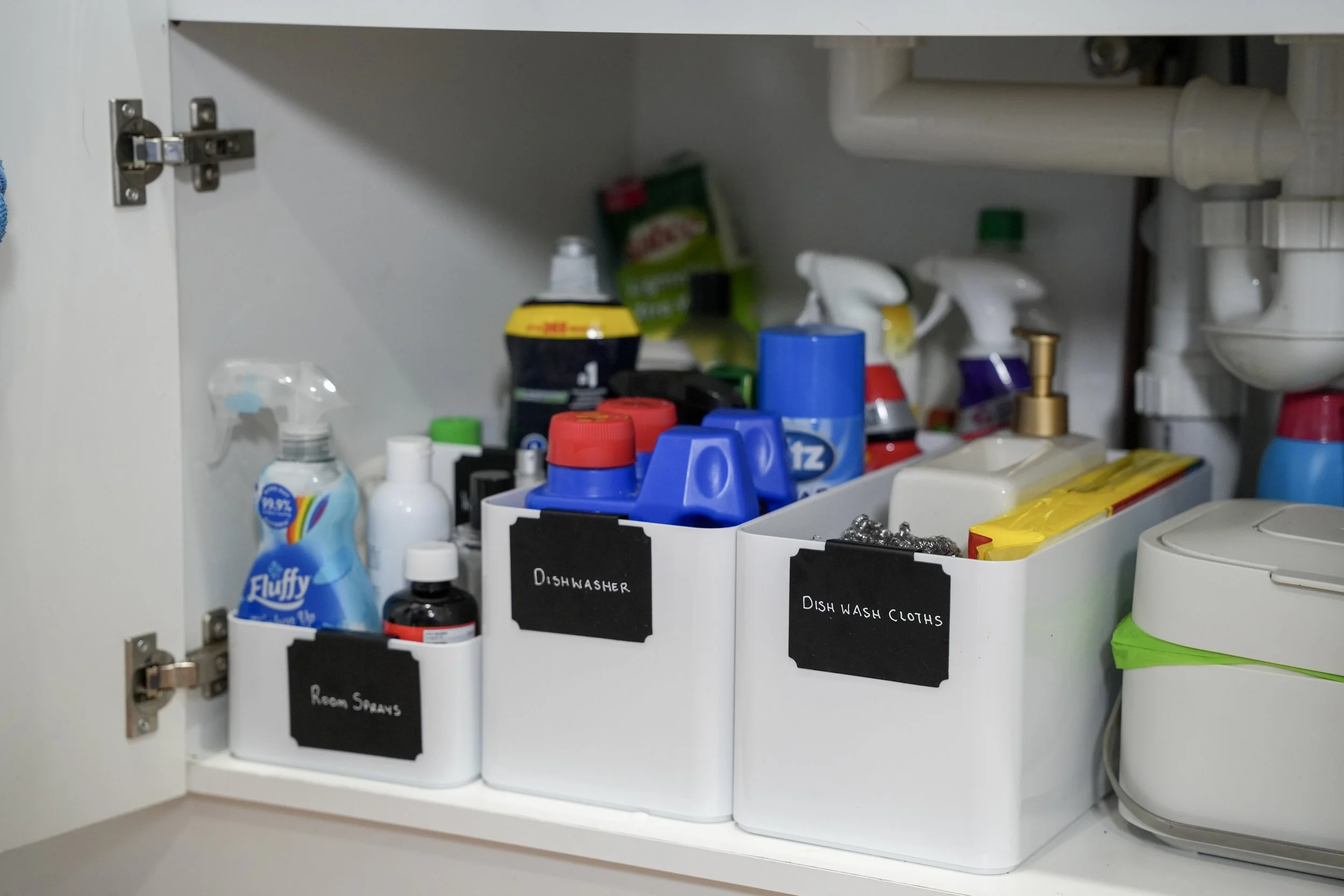 Open kitchen cabinet containing cleaning supplies and dish towels, labeled sections for dishwashers, room sprays, and dishwash cloths.