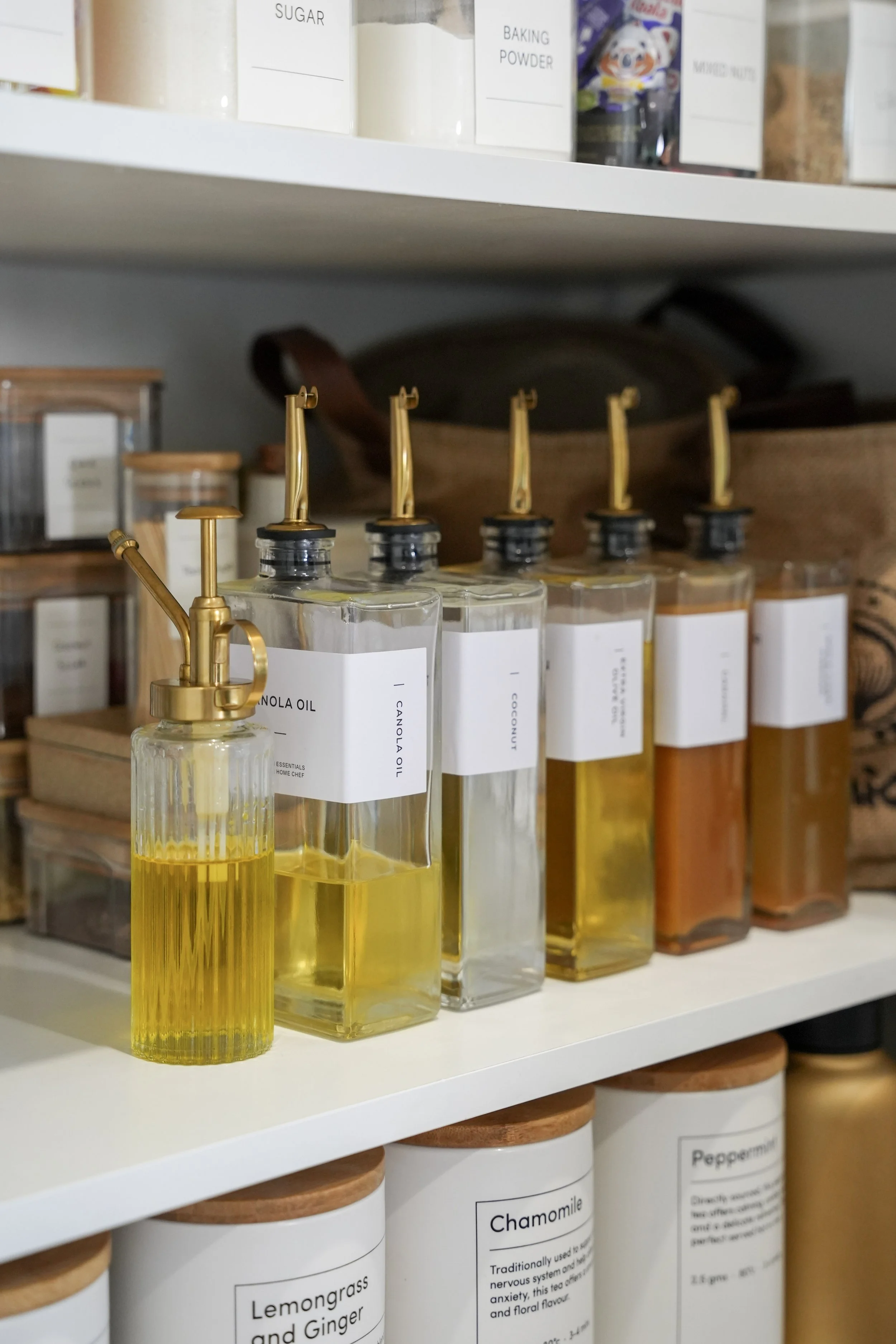 Shelf with glass bottles of cooking oils and jars of herbs and spices, including lemon grass and ginger, chamomile, peppermint, and other seasonings in white containers with wooden lids.