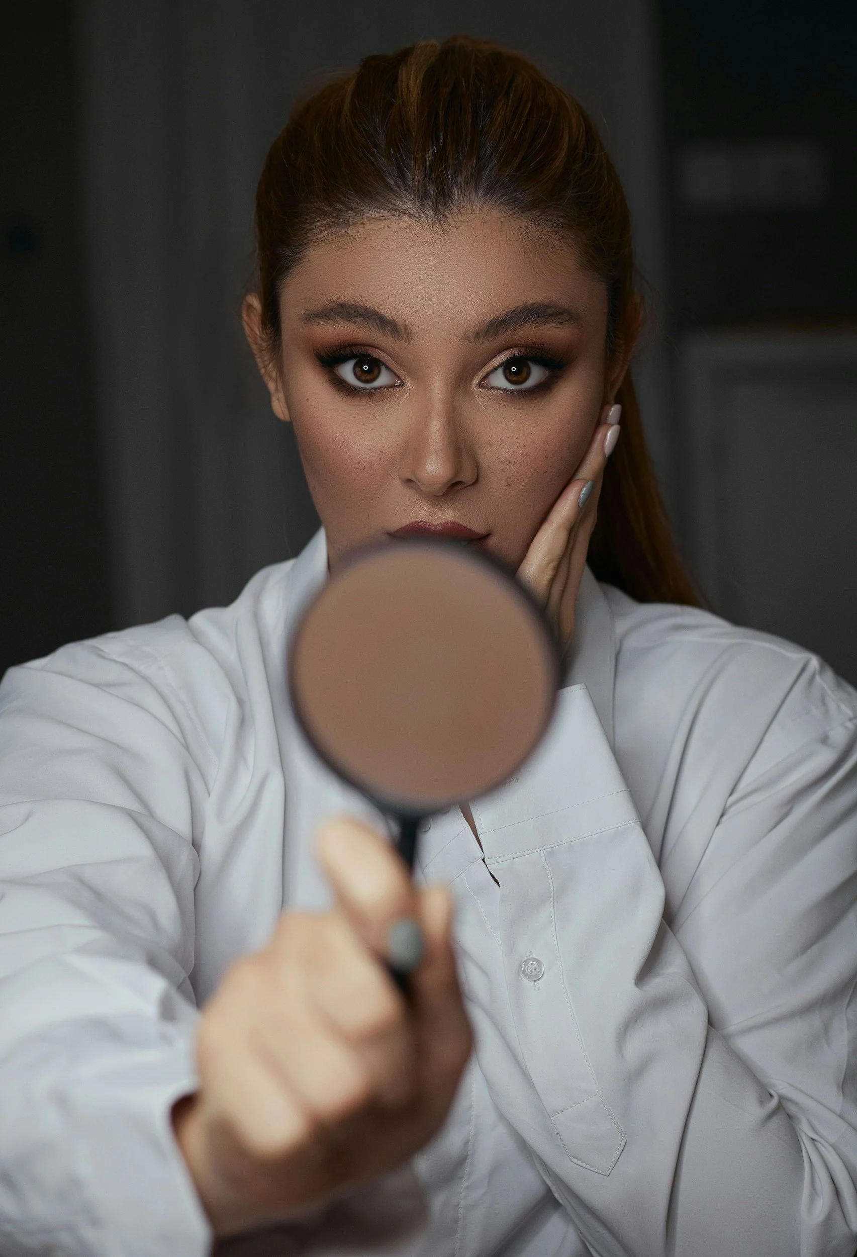 A woman with brown hair and brown eyes wearing a white shirt is holding a makeup brush or applicator closer to the camera, partially obscuring her face, and looking directly at the camera with a neutral expression.