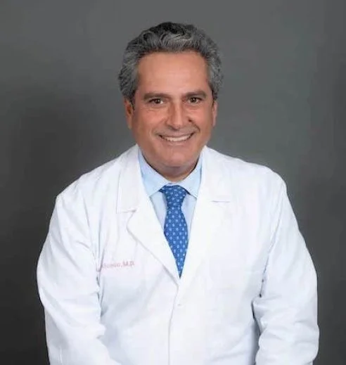 Smiling male doctor in a white coat with blue tie against gray background.