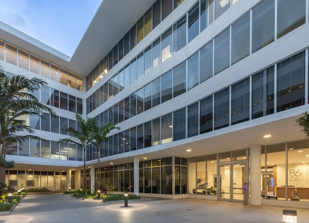 Modern multi-story office building with glass windows, palm trees, and an open courtyard entrance.