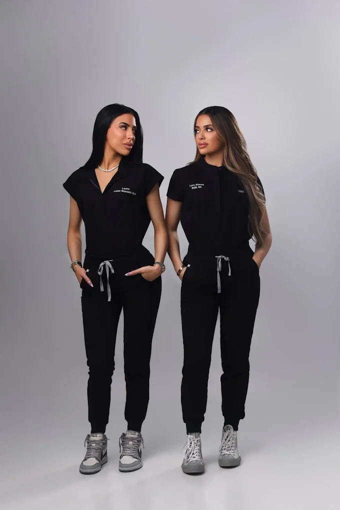 Leann and Lara in matching black uniforms with name tags, standing side by side against a plain gray background, looking at each other with hands in their pockets.