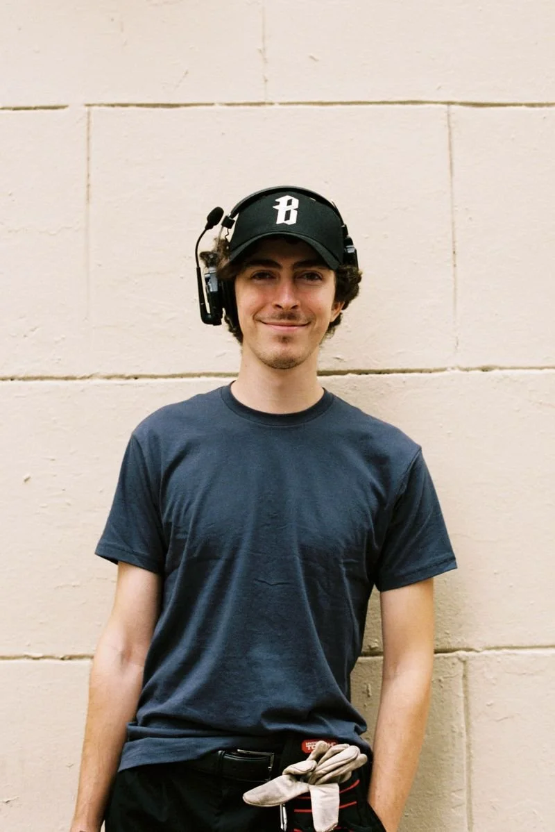 Young man wearing a black baseball cap and headset, standing against a beige brick wall