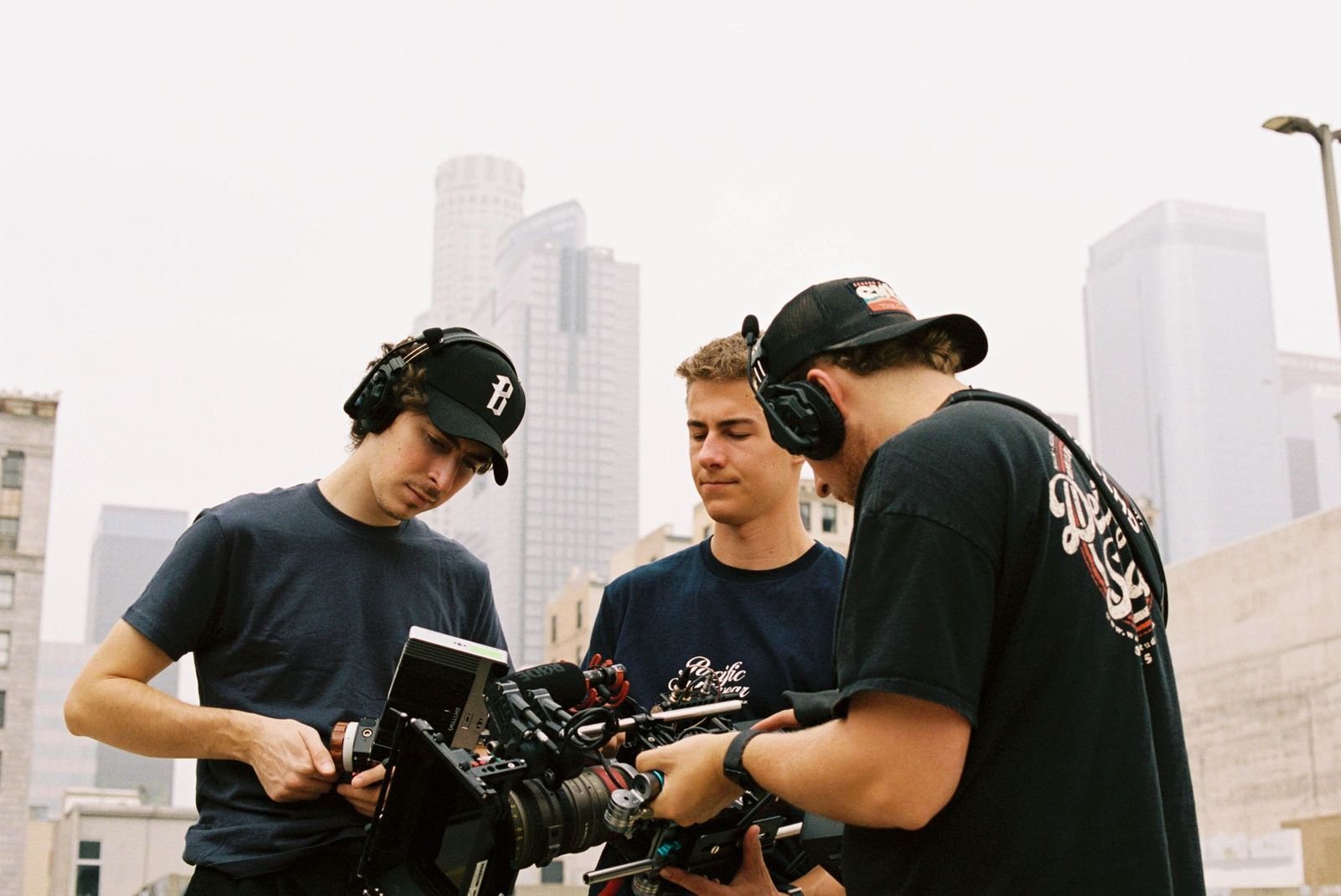 Three filmmakers operate a professional video camera on a city rooftop with skyscrapers in the background.