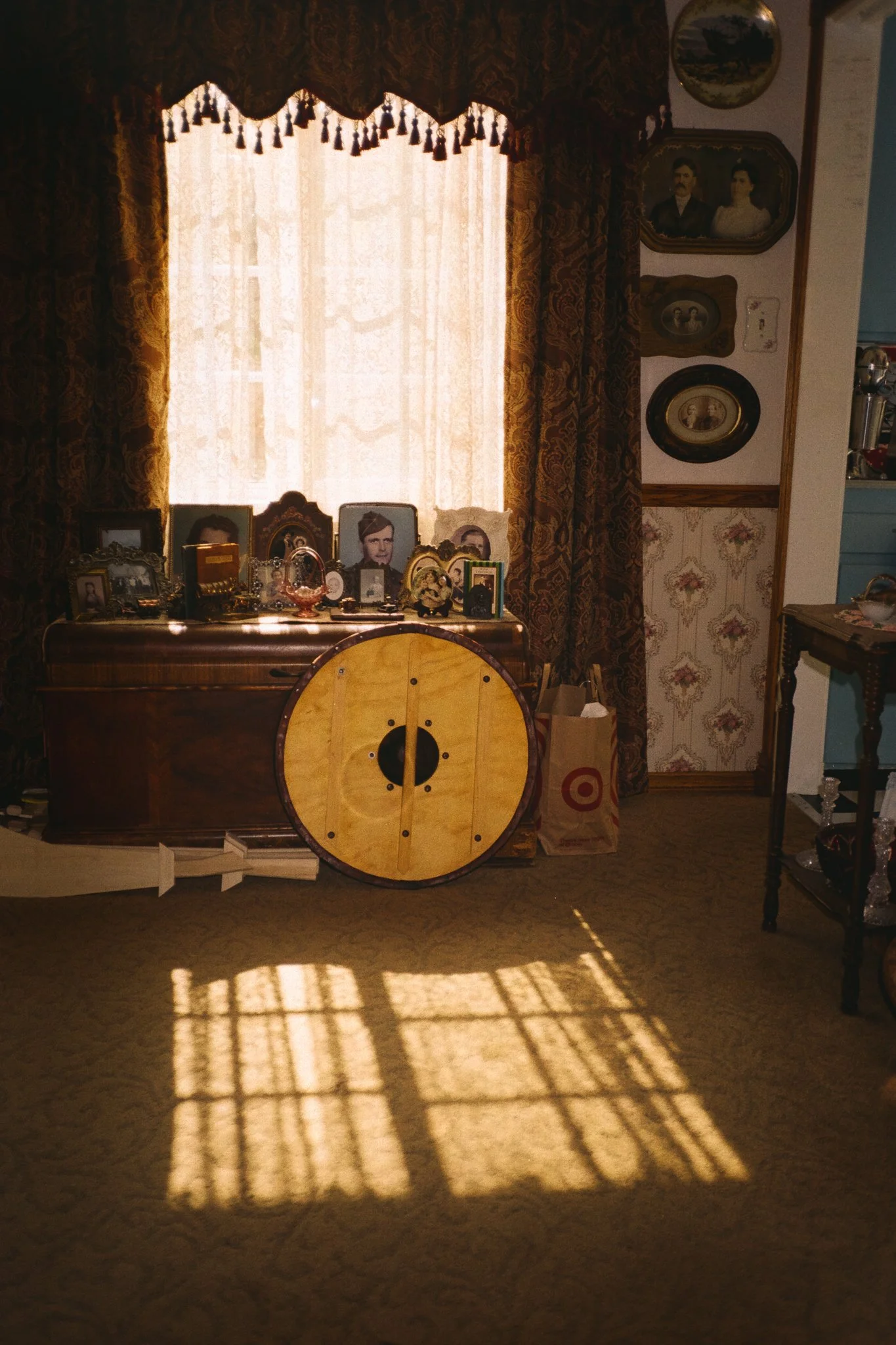 Sunlit vintage living room with family photographs, patterned curtains, antique furniture, and a large round wooden shield leaning against a dresser.