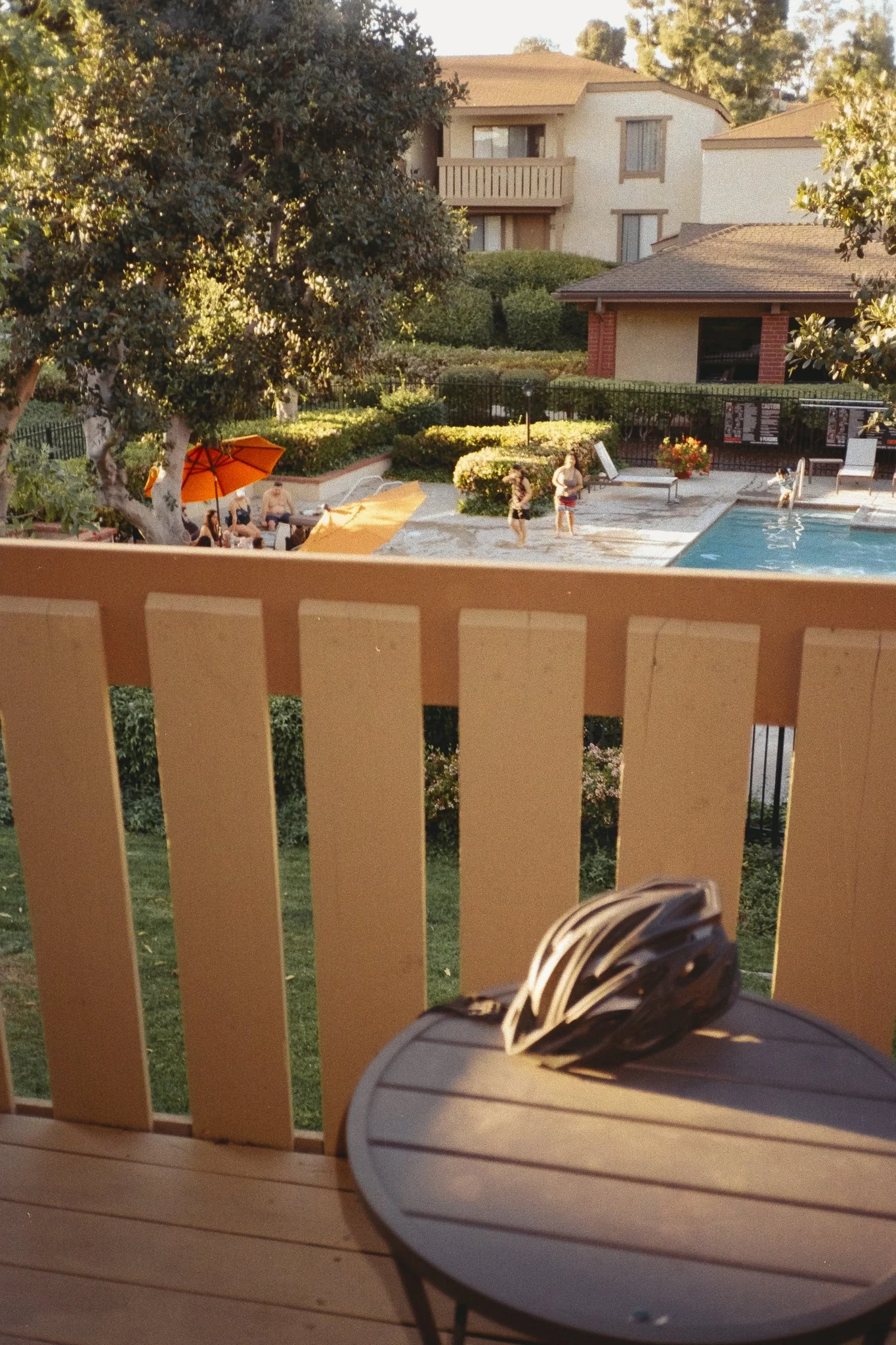 Apartment courtyard pool with people, lounge chairs, orange umbrellas, lush landscaping, and a balcony railing in the foreground