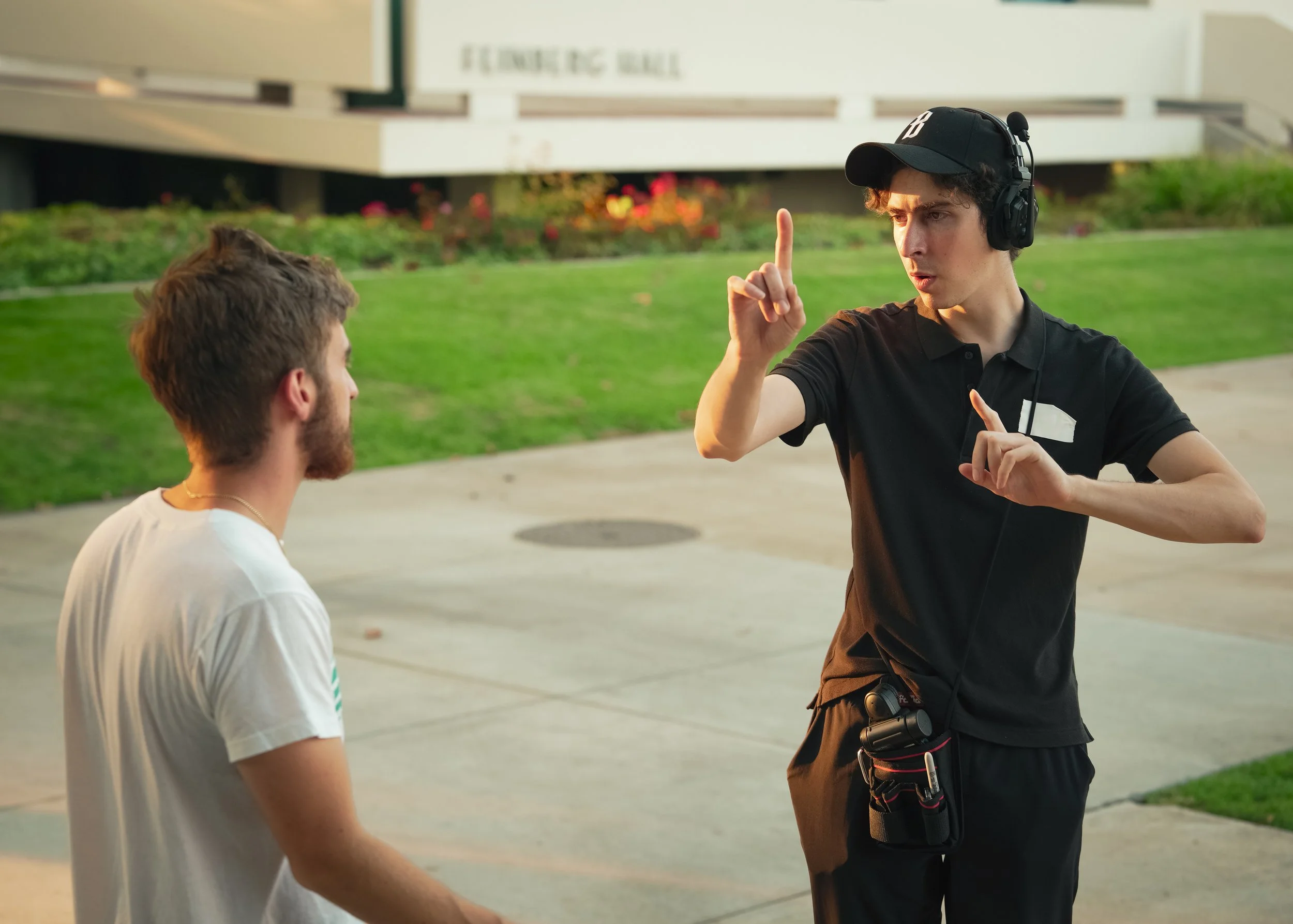 A sign language interpreter wearing a headset gestures while communicating with a man outdoors.
