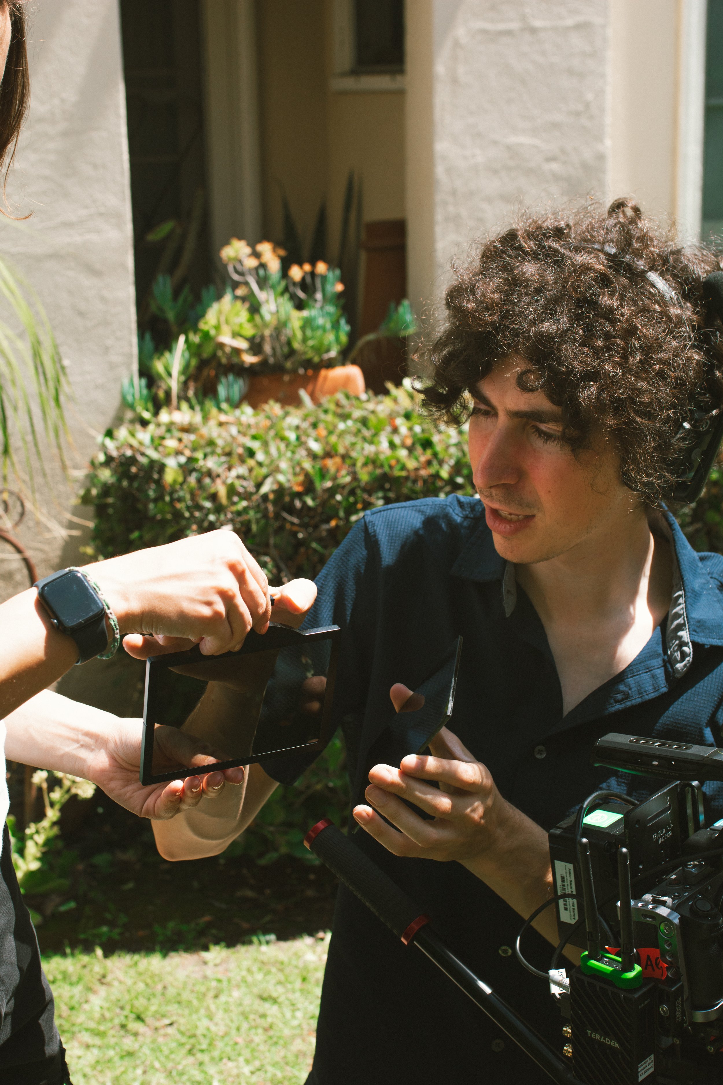 Film crew member with curly hair holding a camera slate beside professional camera equipment outdoors