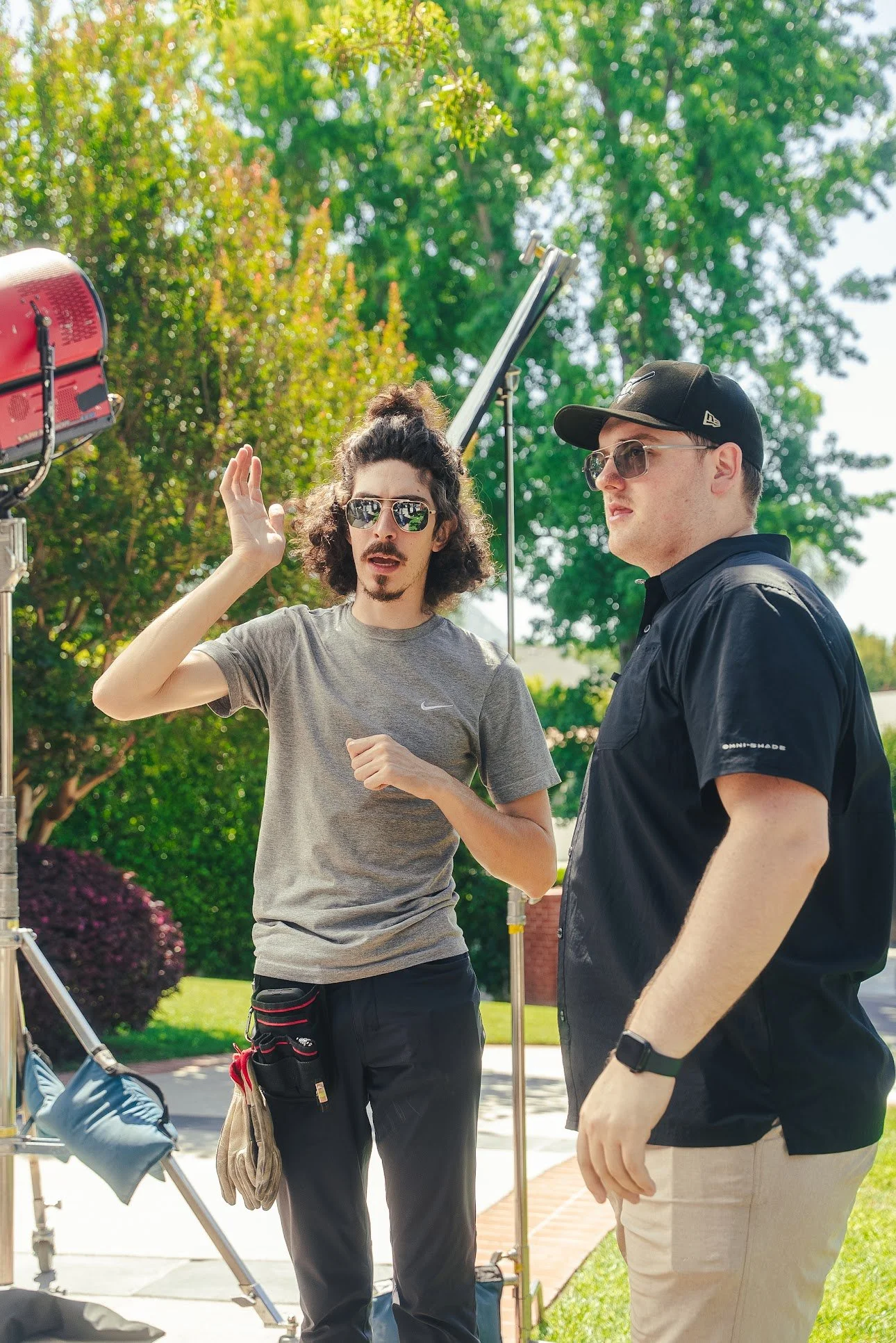 Two men wearing sunglasses stand outdoors beside a film production setup, with one gesturing and a studio light visible behind them.