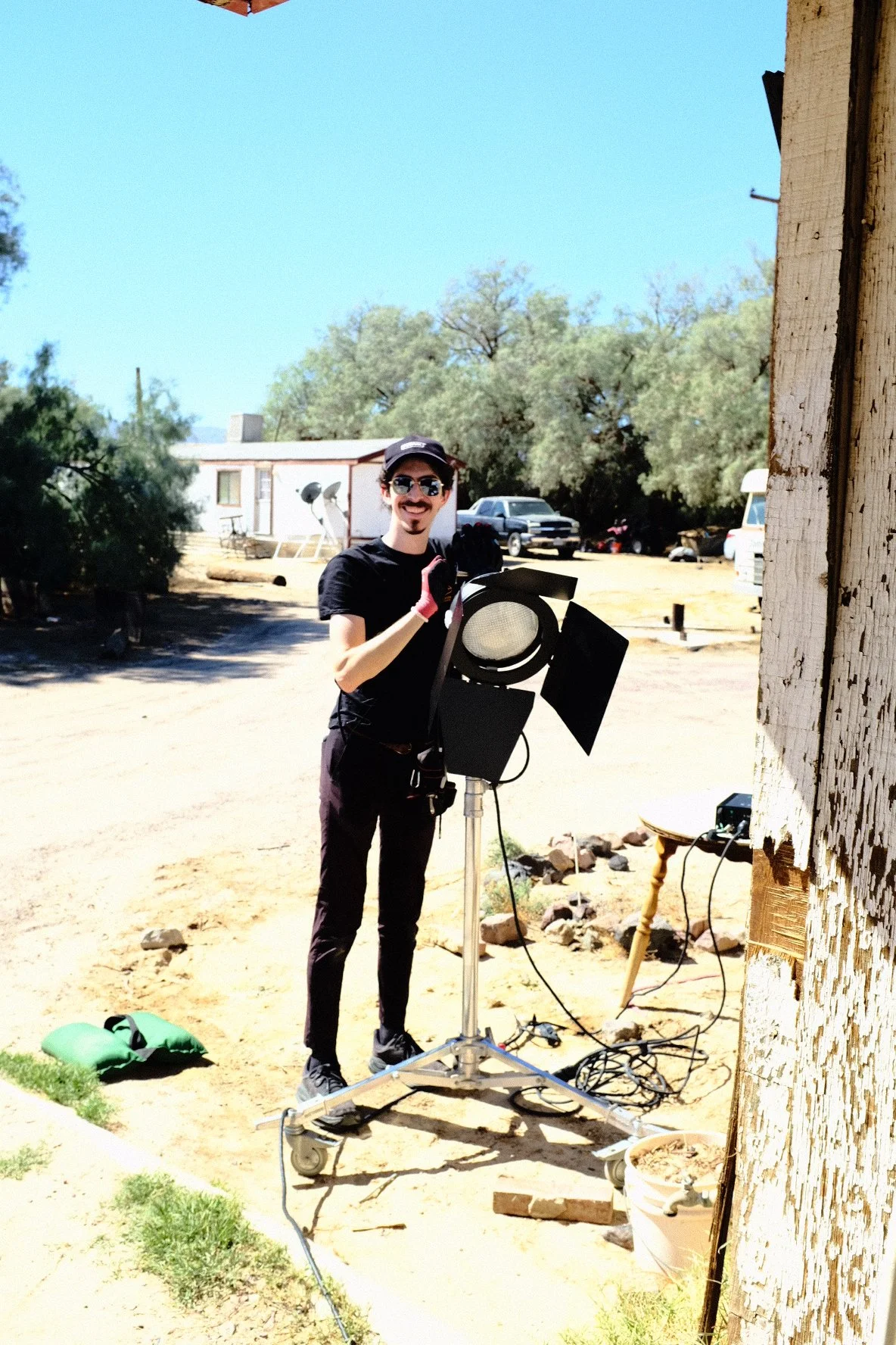 Film crew member wearing sunglasses and a black cap stands beside a studio light on a wheeled stand outdoors.