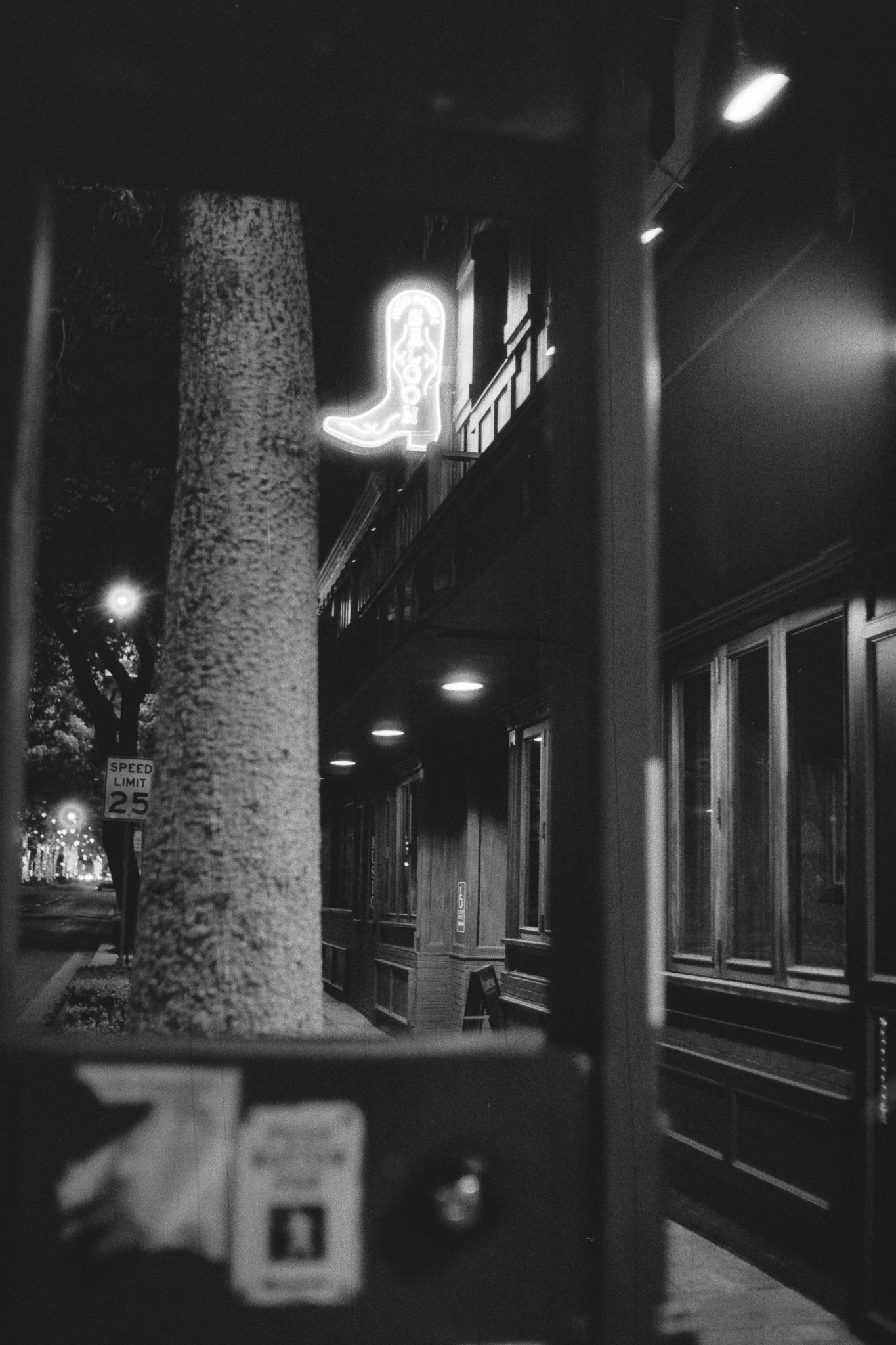 Black-and-white nighttime street scene with a neon cowboy boot sign outside a building, sidewalk, trees, and a 25 mph speed limit sign.