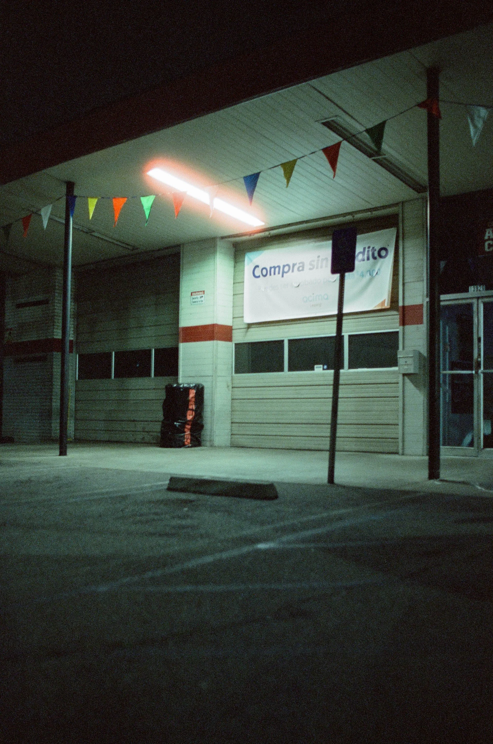 Nighttime view of a brightly lit convenience store with colorful pennant flags and a Portuguese sale sign