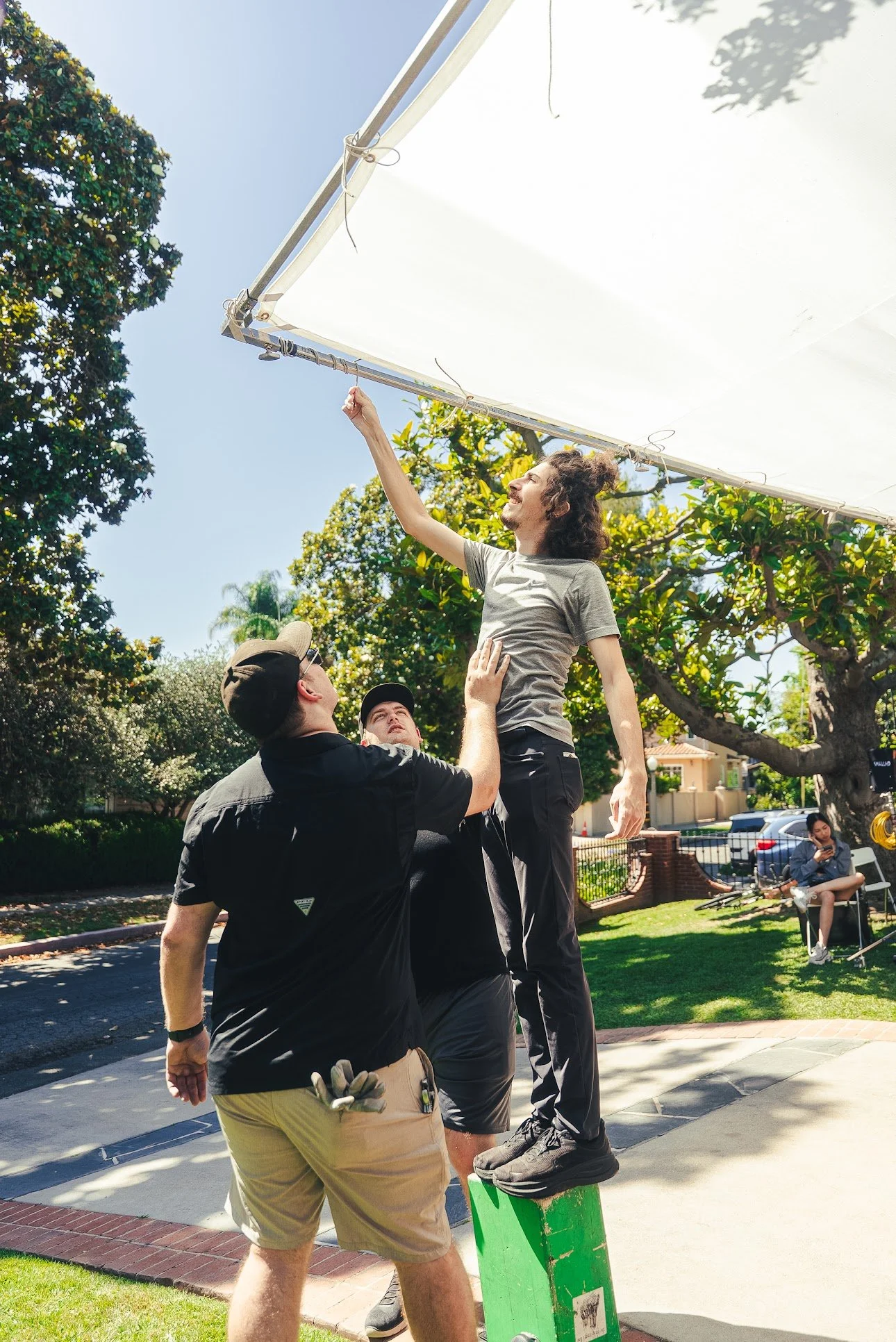 Film crew lifting a man to adjust a large outdoor reflector in a sunny residential yard